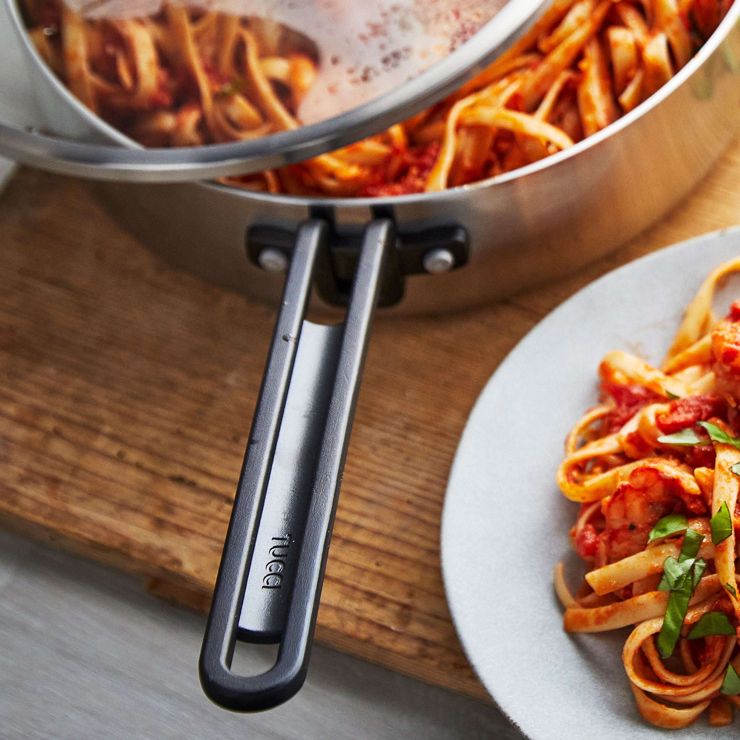 Stainless steel nonstick pan with tomato pasta and basil on a wooden surface.