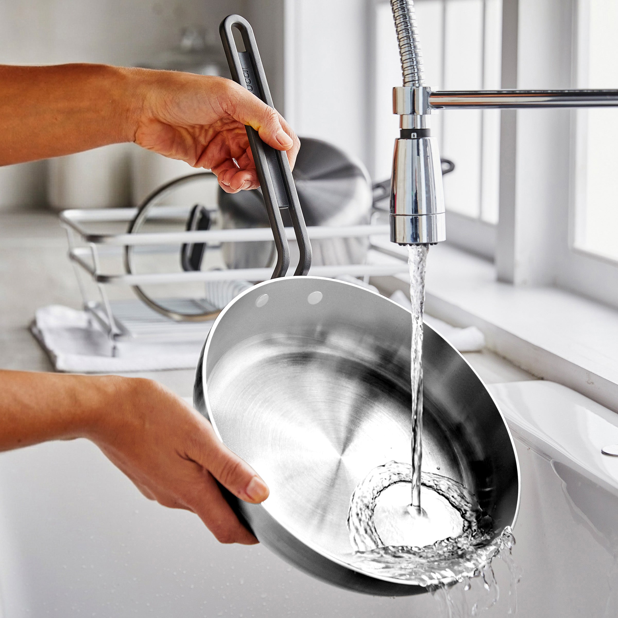 Rinsing a stainless steel ceramic nonstick pan under running water at a kitchen sink.