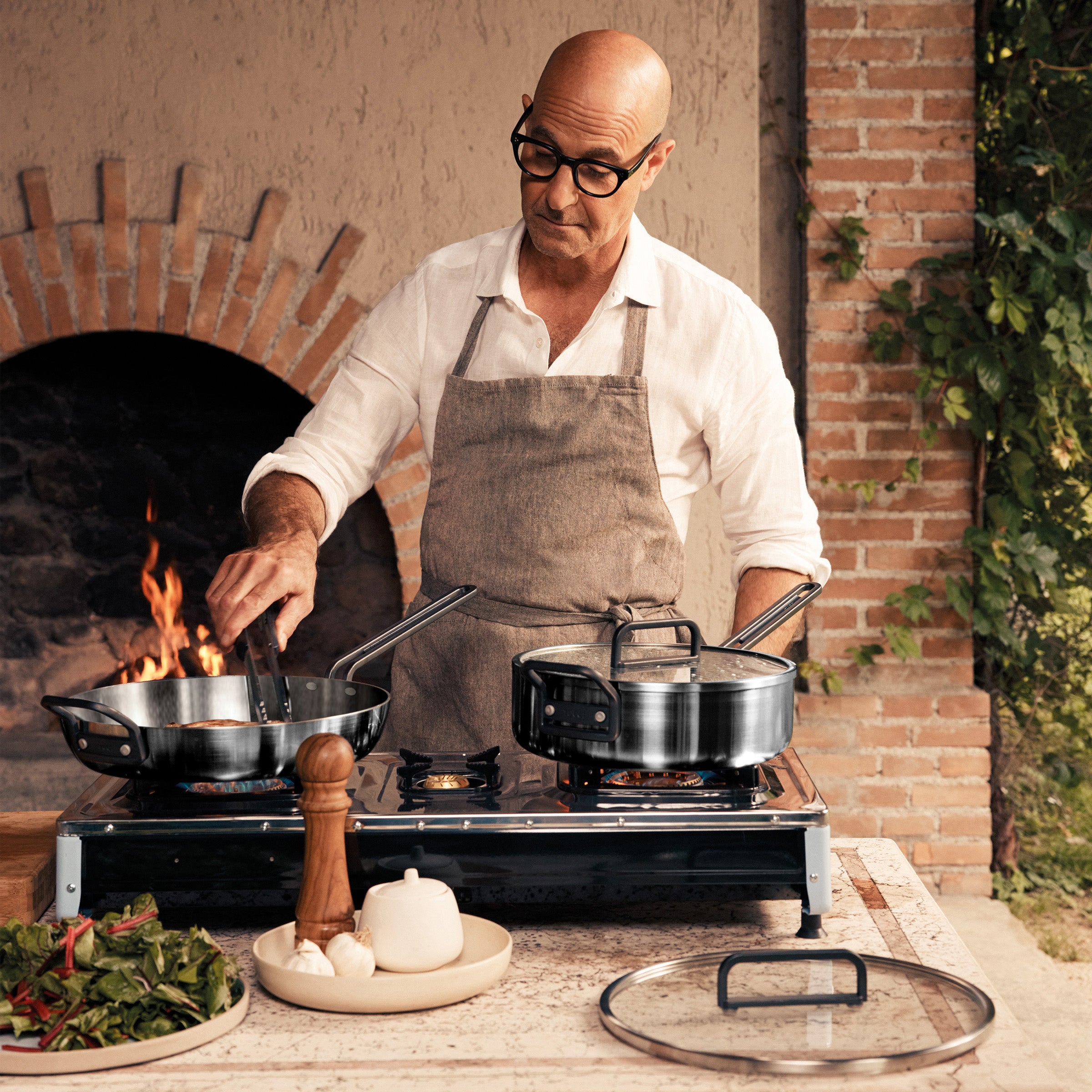 Man in apron cooks vegetables outdoors, stirring a pan from the Stanley Tucci 9-piece cookware set with glass lids.