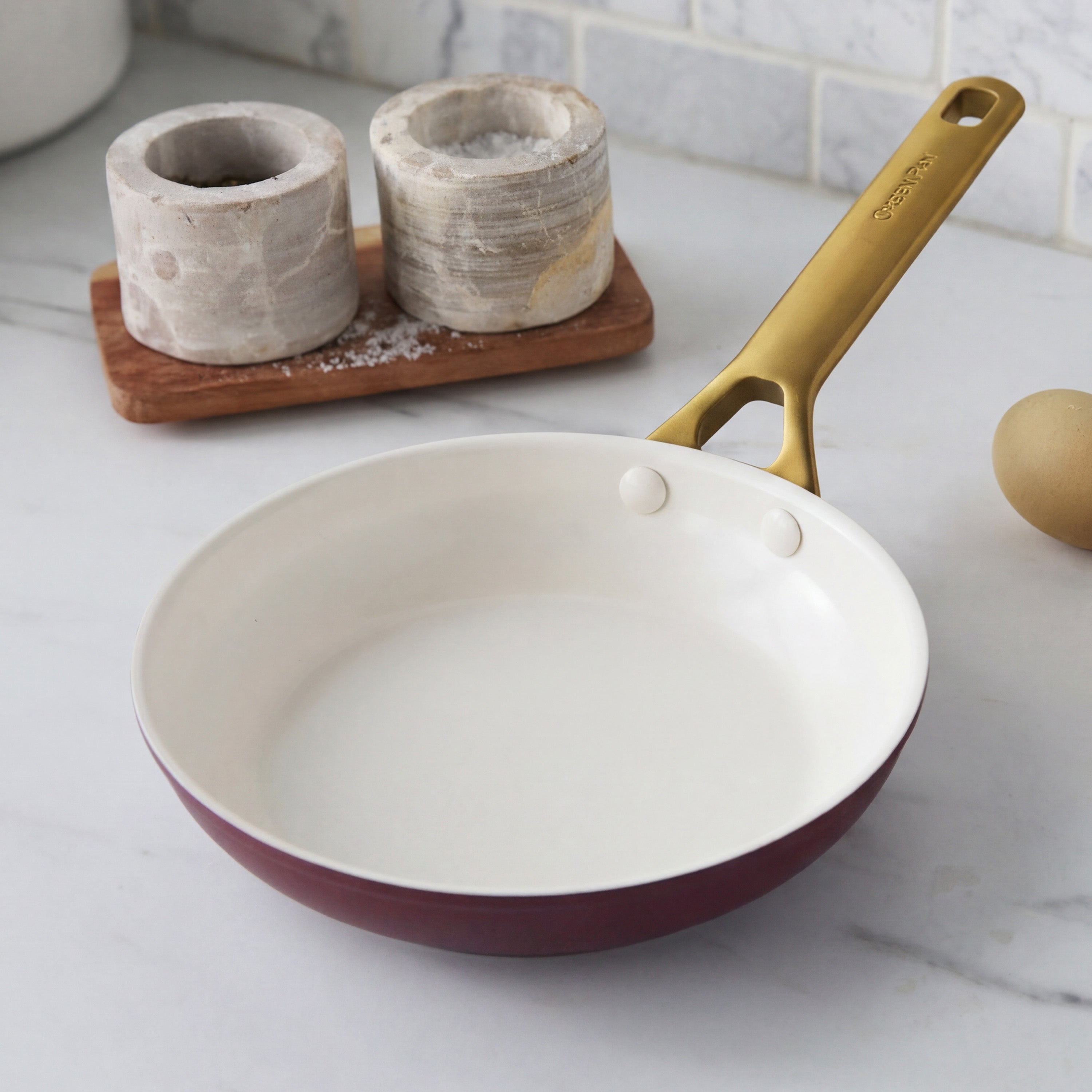 Bordeaux ceramic frypan with a gold handle on marble, next to salt holders and an egg.