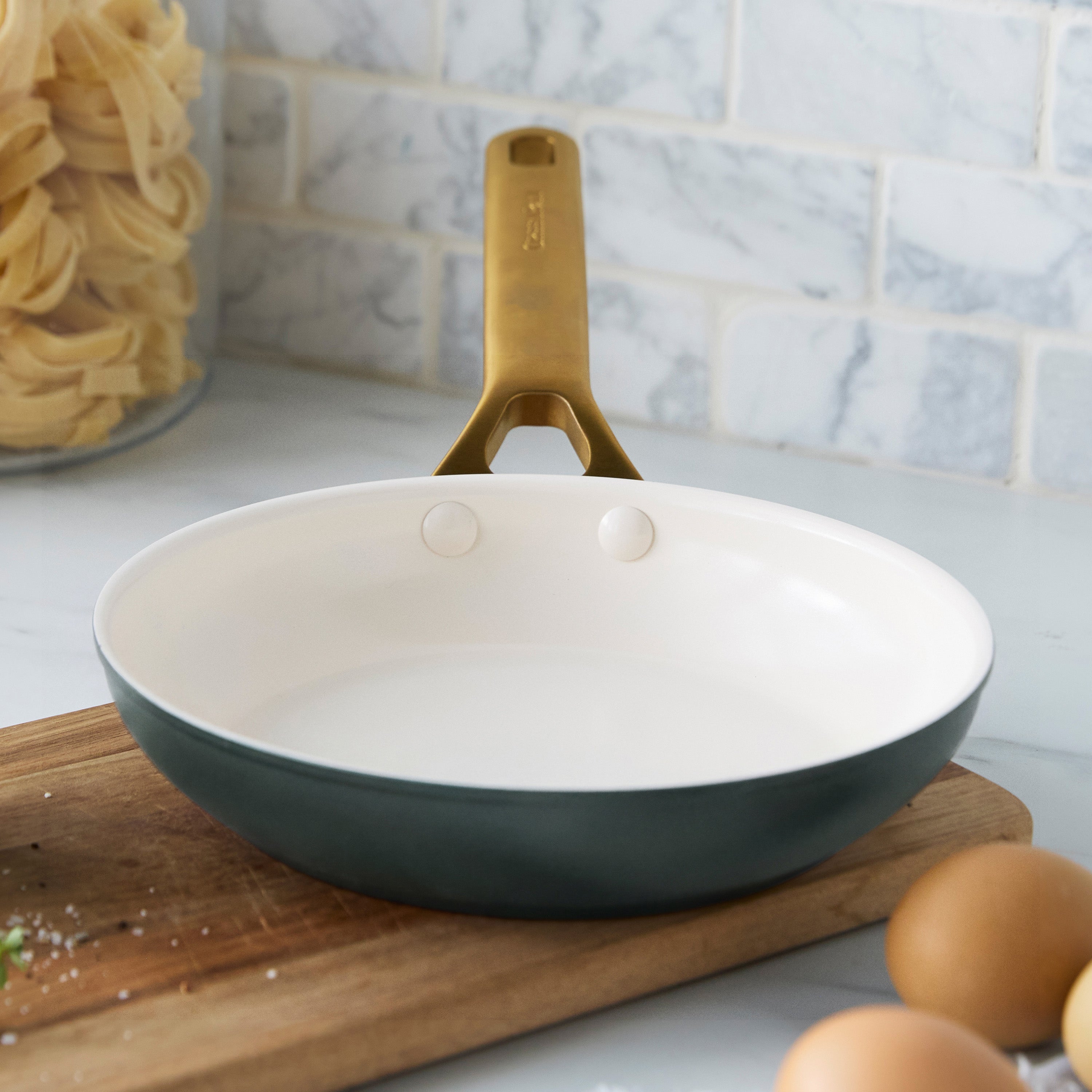 Spruce green ceramic nonstick 8-inch frypan with gold handle on a counter next to eggs, pasta, and a cutting board.