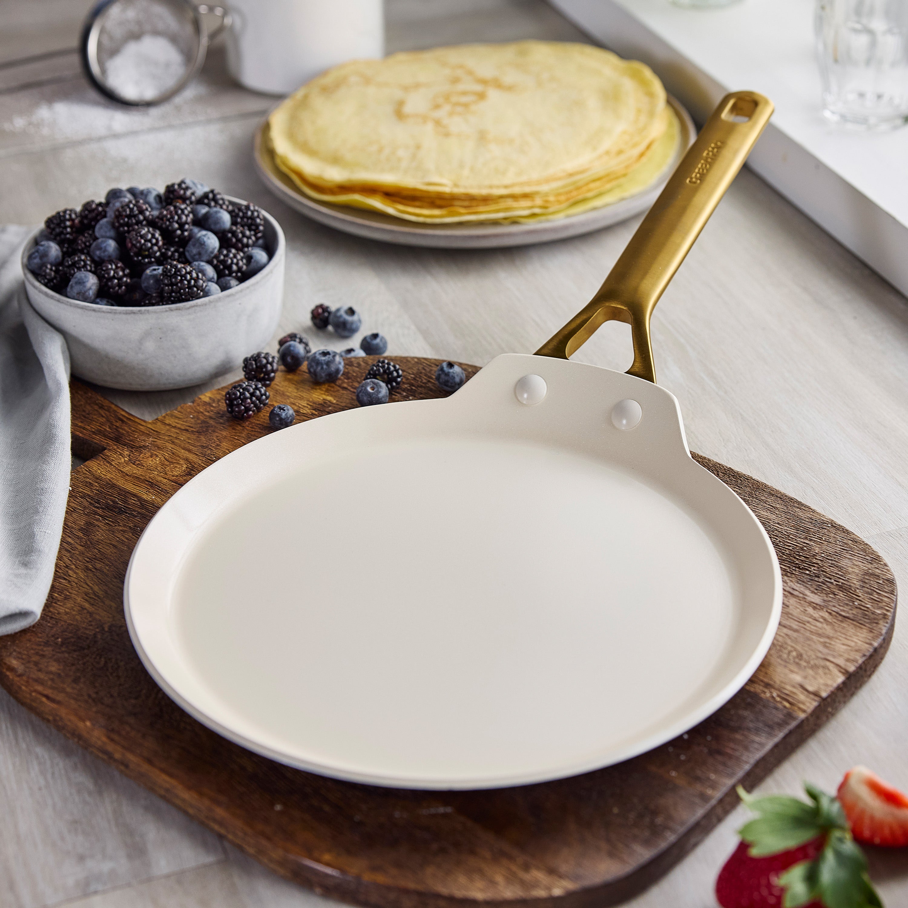 Bordeaux ceramic nonstick crepe pan with gold handle on wood board; berries and stacked crepes in background.