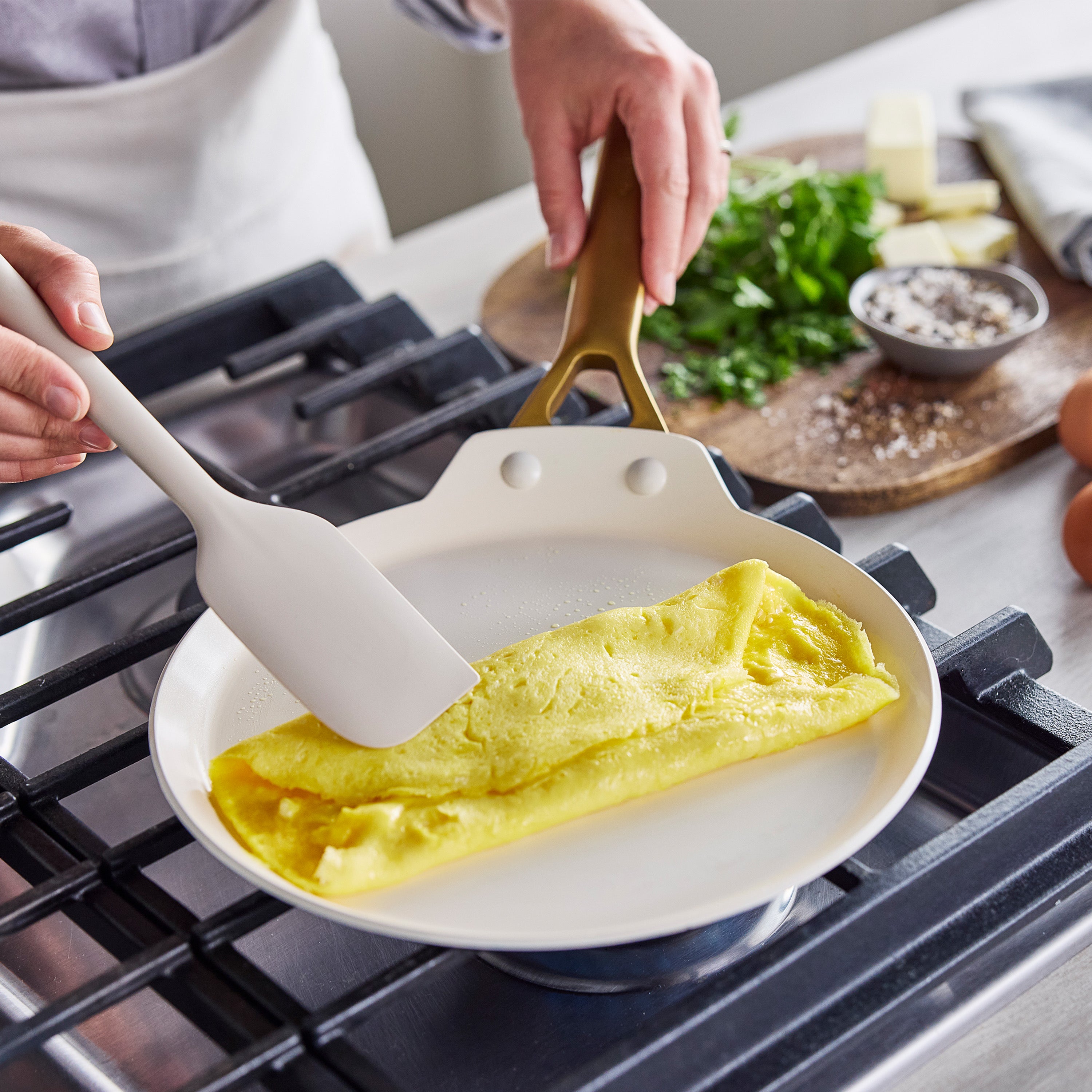 Yellow omelet being folded in a Bordeaux red ceramic nonstick crepe pan, with eggs and herbs on a wooden surface nearby.