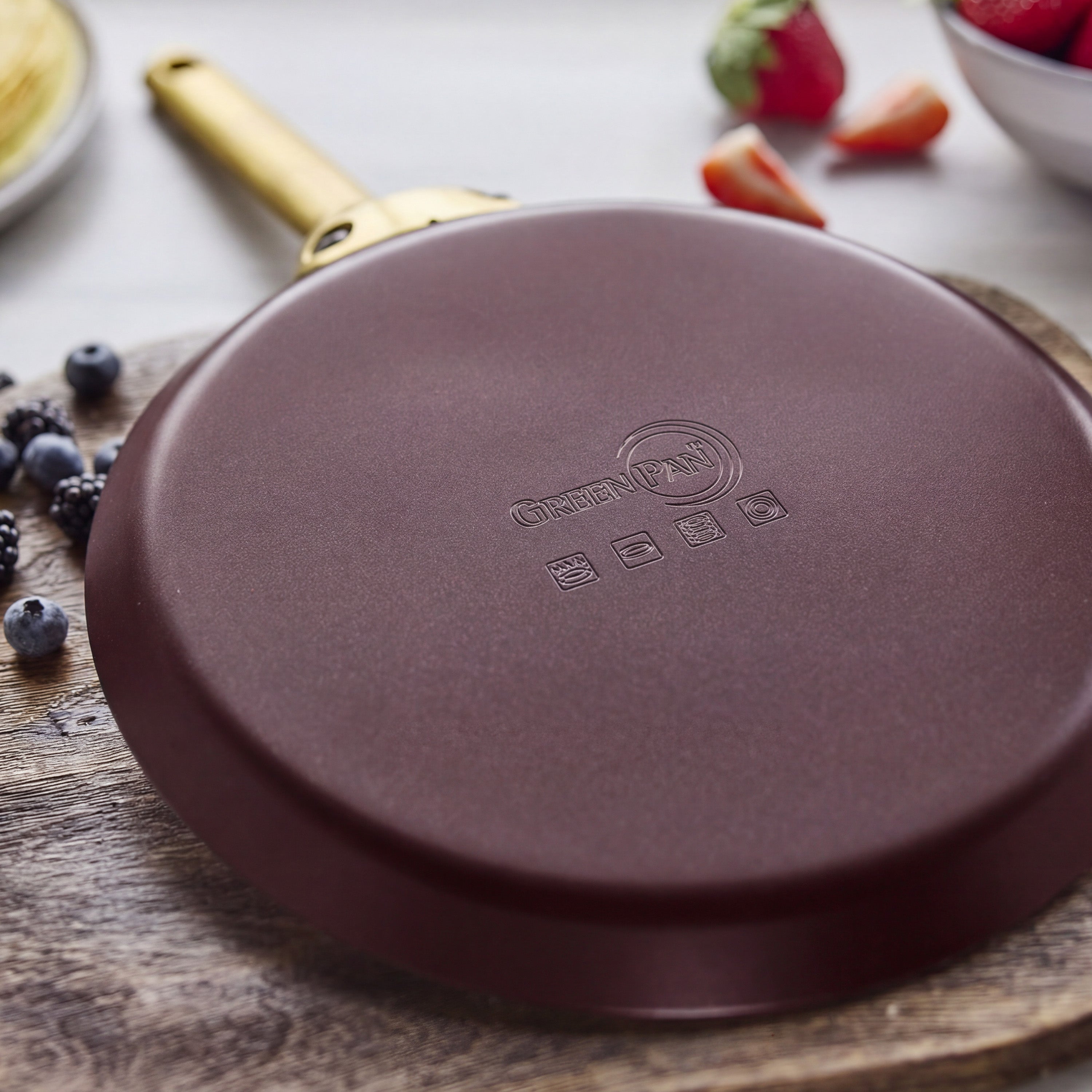 Red ceramic nonstick crepe pan, 10-inch, shown upside down on a wooden board with berries nearby and a gold handle visible.