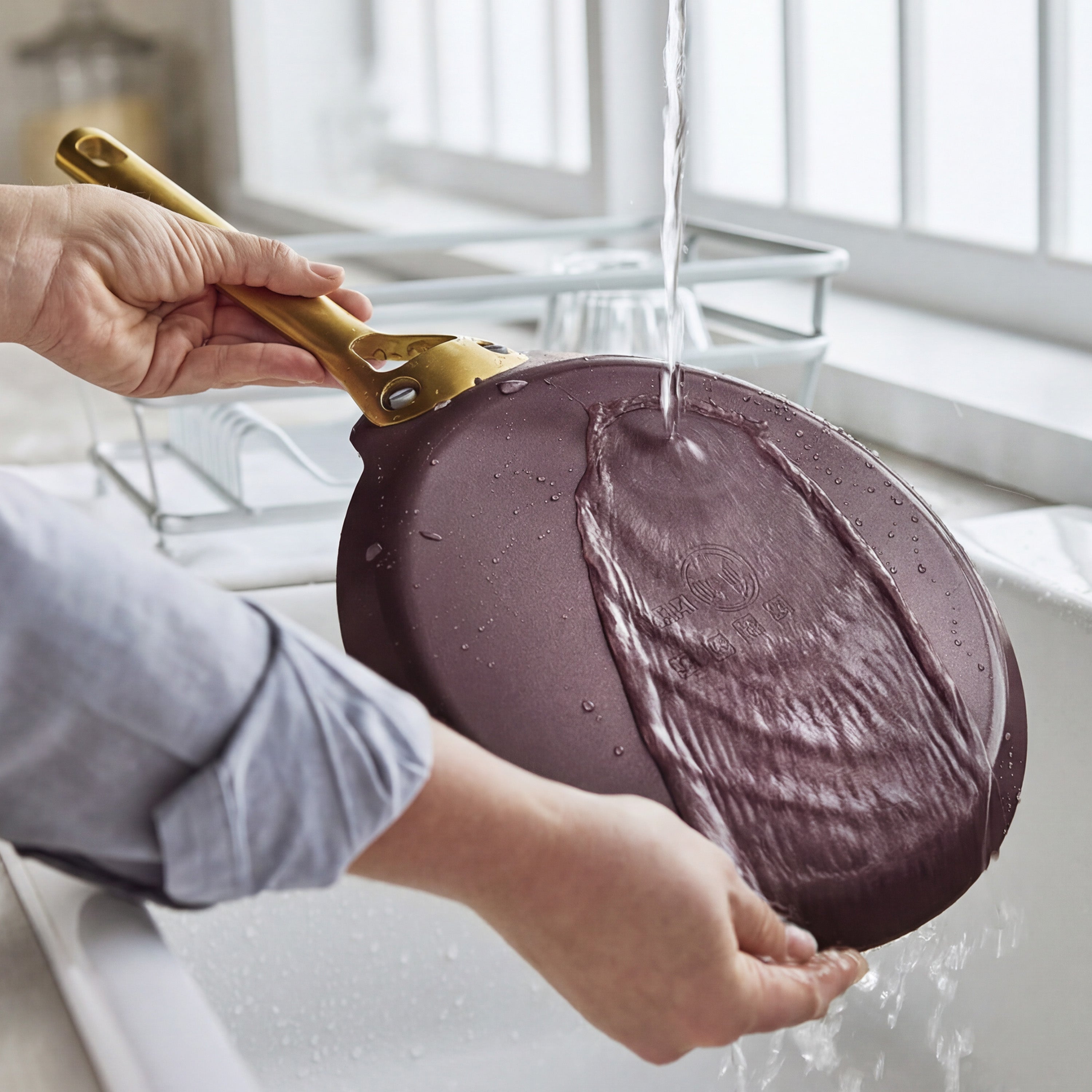 A Bordeaux red ceramic nonstick crepe pan is rinsed under running water in a kitchen sink by a window.