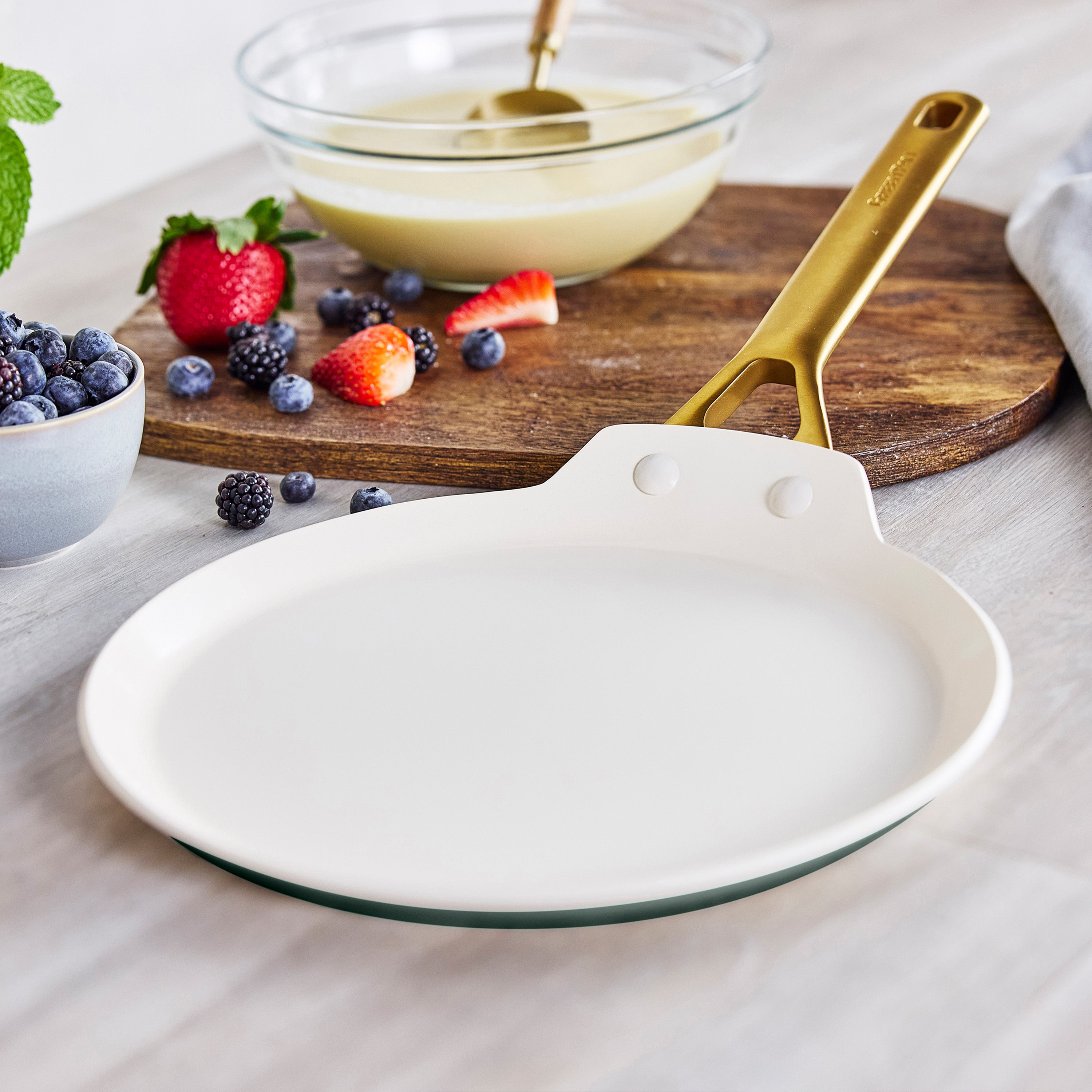 Spruce green ceramic nonstick crepe pan with gold handle on table beside berries, batter bowl, and wooden cutting board.