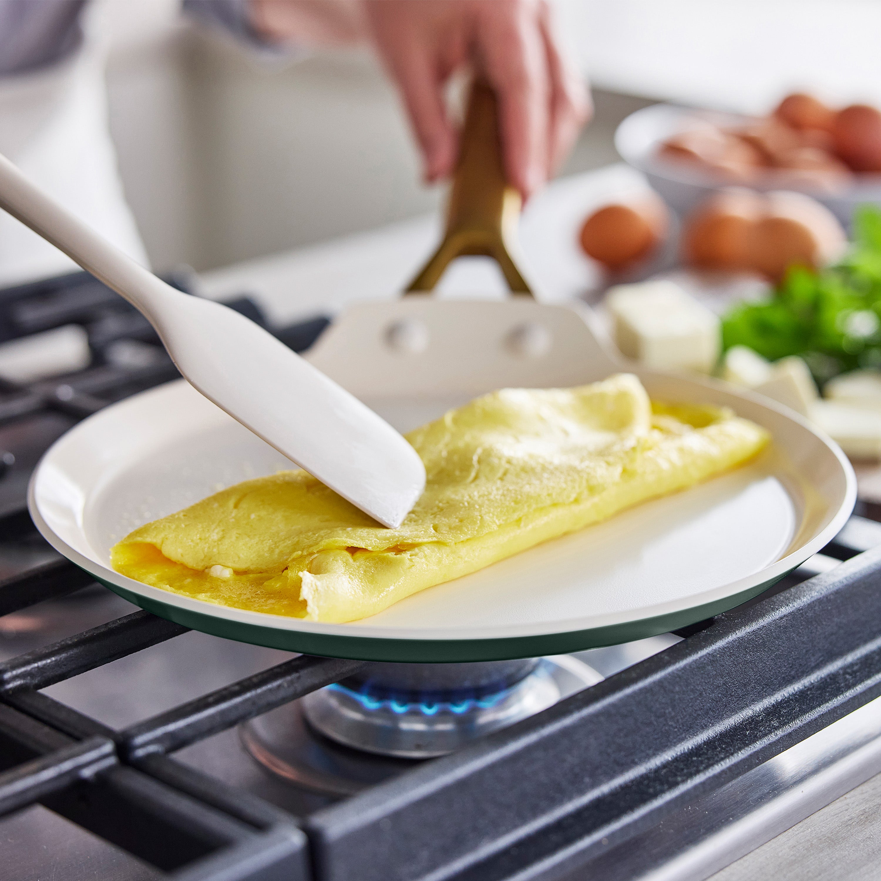 Folding an omelet in a black ceramic nonstick crepe pan with a white spatula, eggs and fresh herbs on the side.
