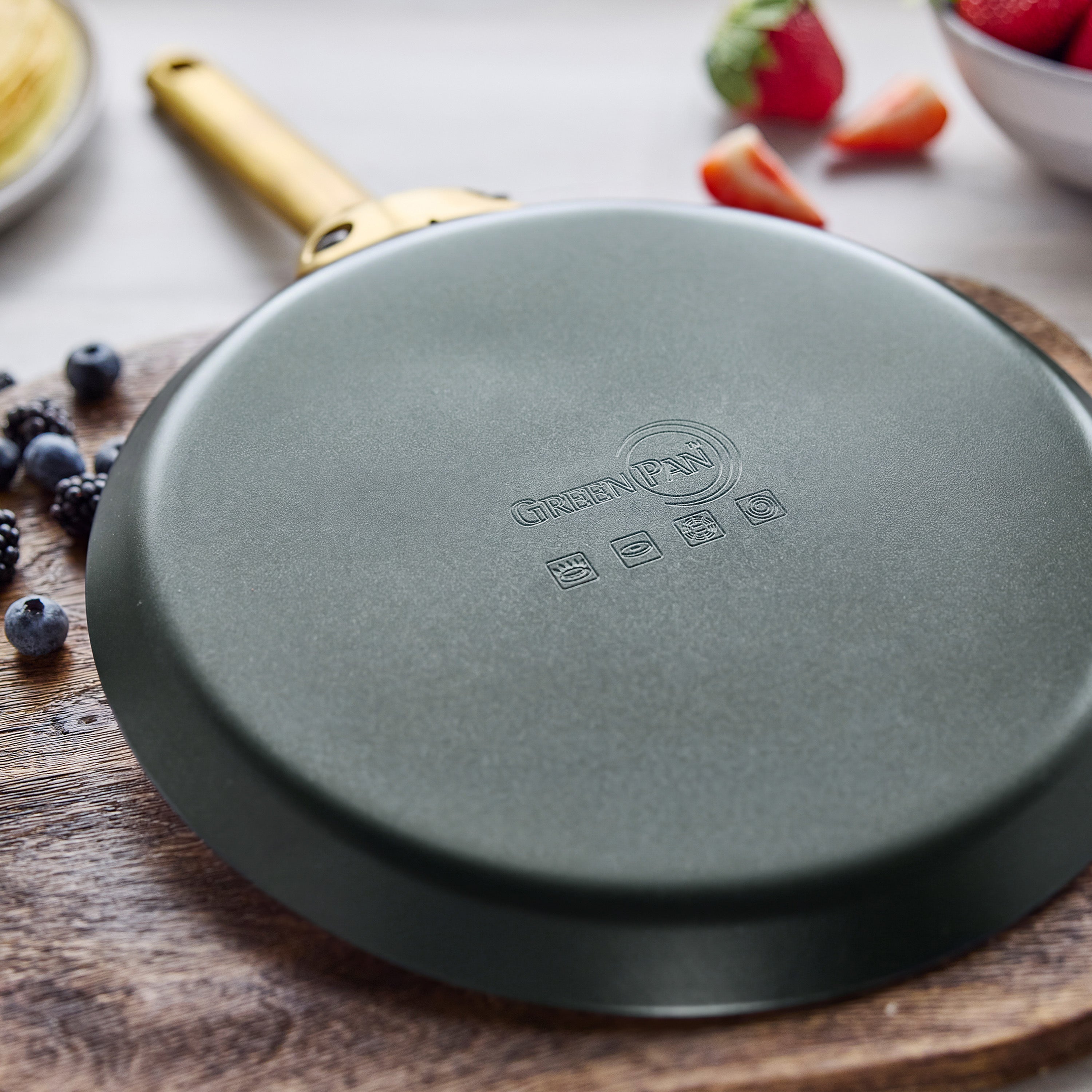 Ceramic nonstick crepe pan in spruce green on a board, with out-of-focus berries and a fruit bowl in the background.