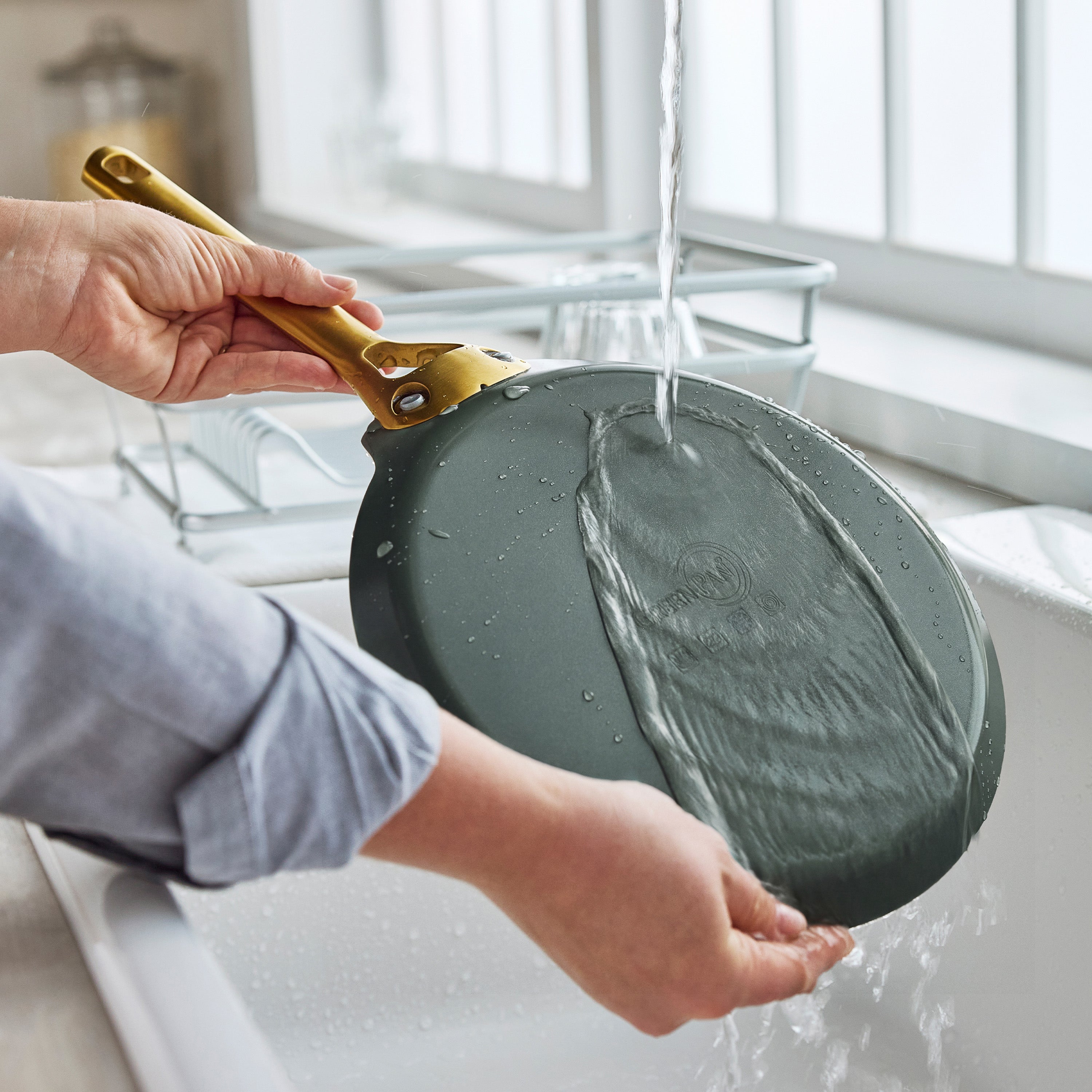 A hand rinsing a black ceramic nonstick crepe pan under running water by a kitchen sink with a drying rack nearby.