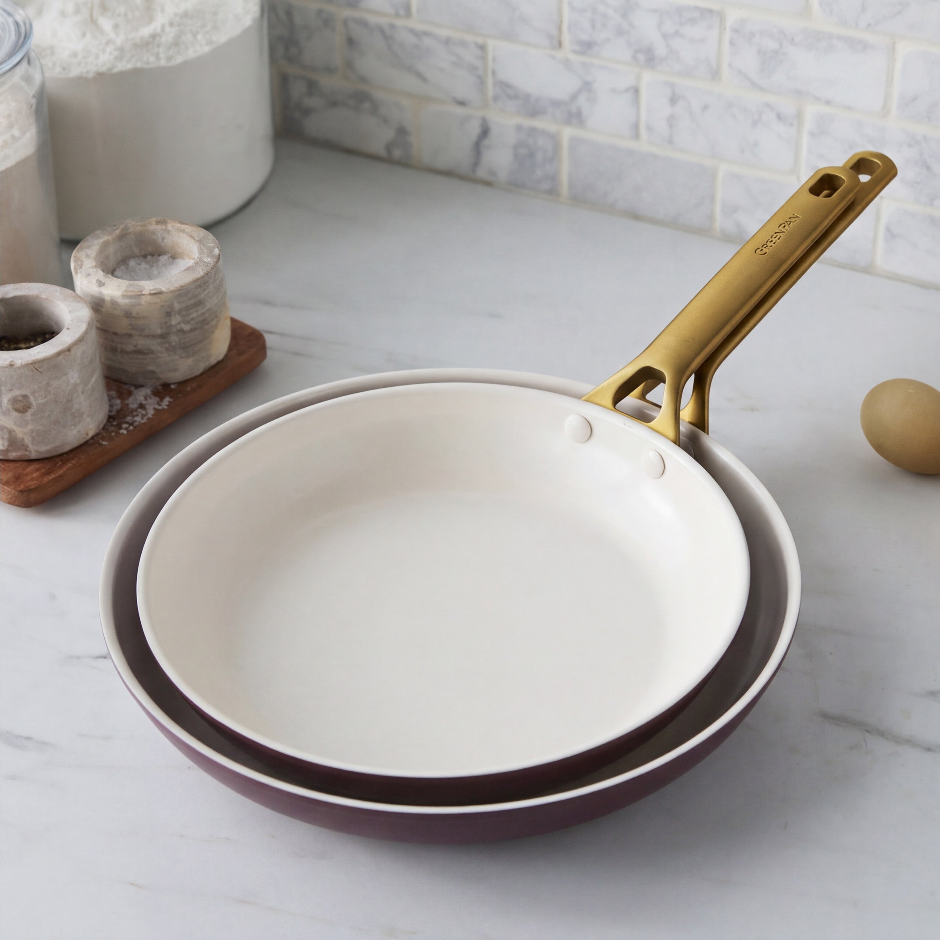 Two Bordeaux ceramic frypans with gold handles on marble, beside salt cellars and a brown egg. Set of two.