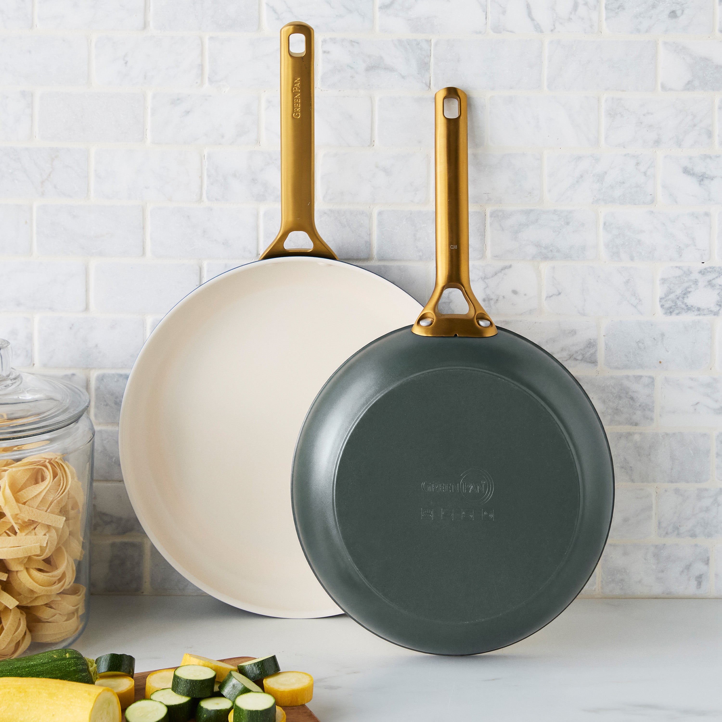 Two spruce green ceramic frypans with gold handles beside pasta and zucchini on a kitchen counter near white tile backsplash.