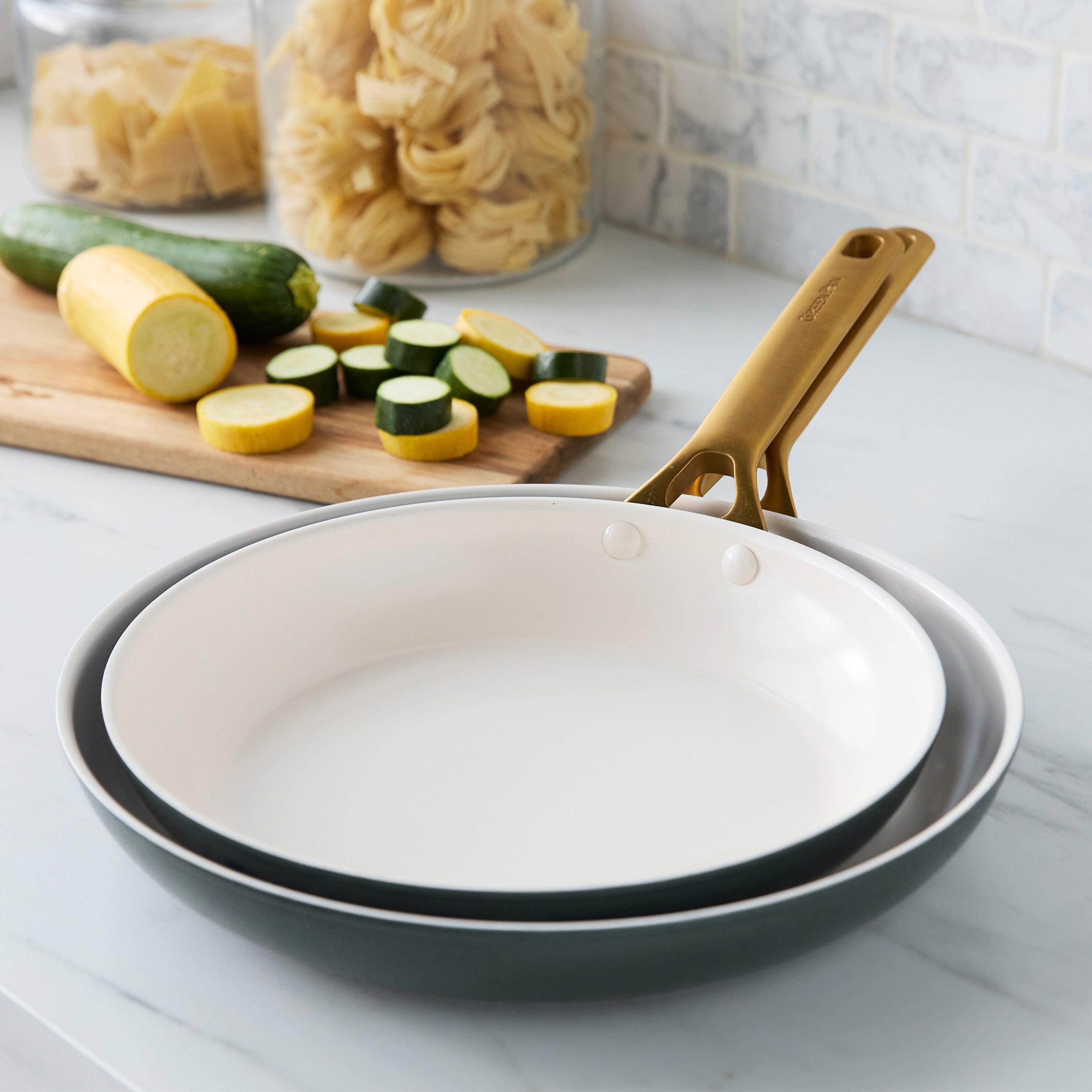 Set of two spruce green ceramic nonstick frypans on a counter, with zucchini, squash, and pasta in the background.