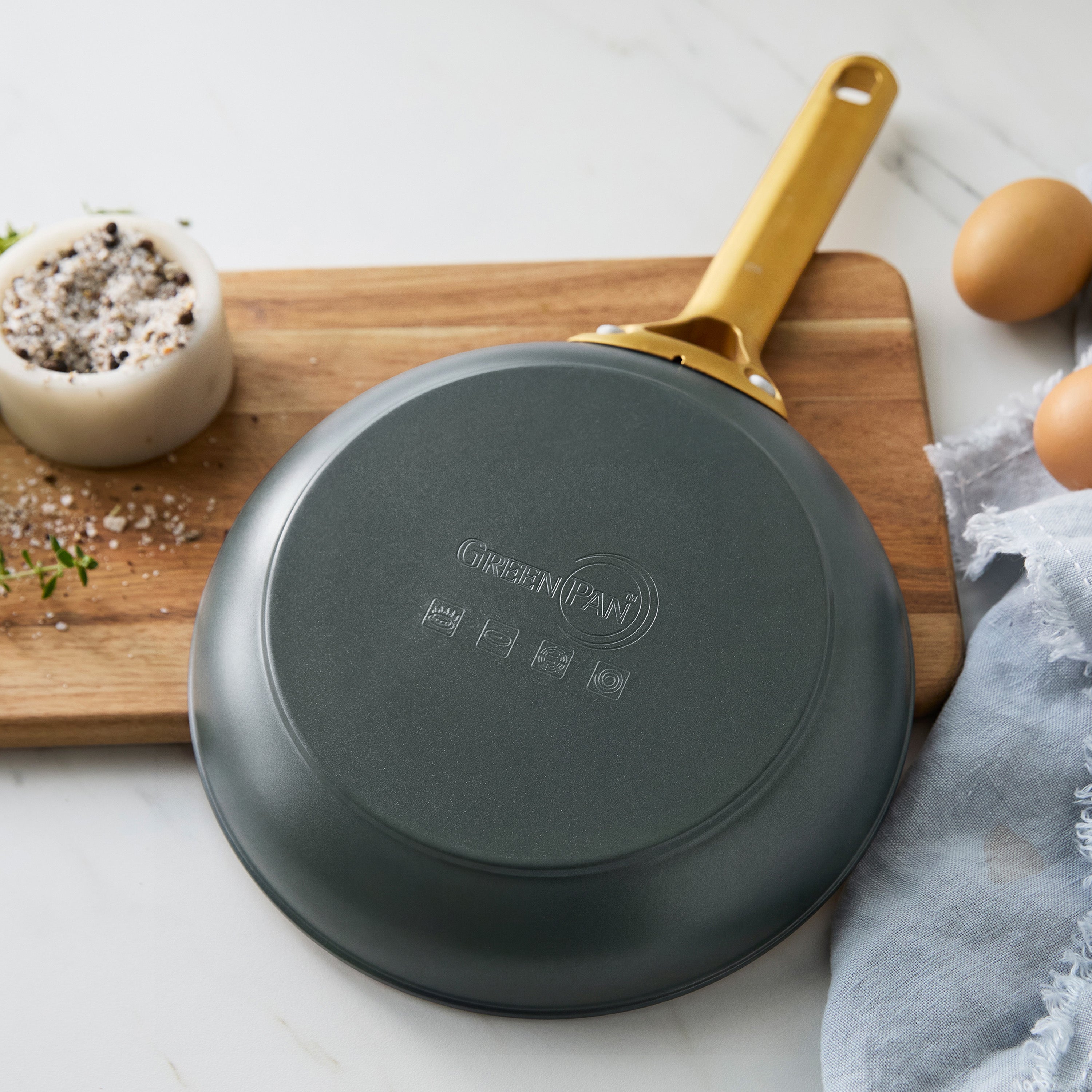 Two spruce green ceramic nonstick frypans, 10″ and 12″, shown upside-down on a board with eggs, salt, and herbs on a white counter.