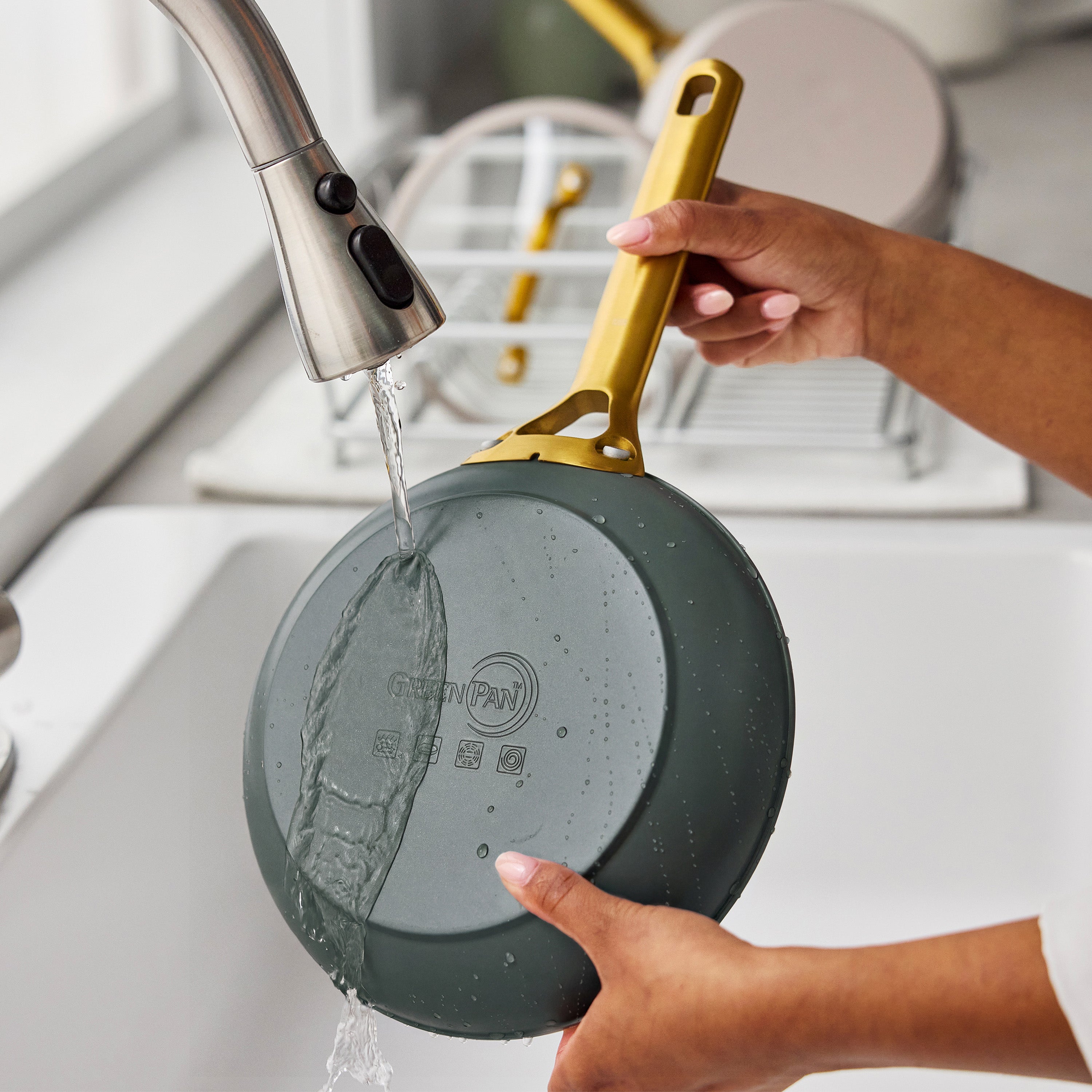 A yellow-handled ceramic nonstick frypan being rinsed under a kitchen faucet in a white sink.