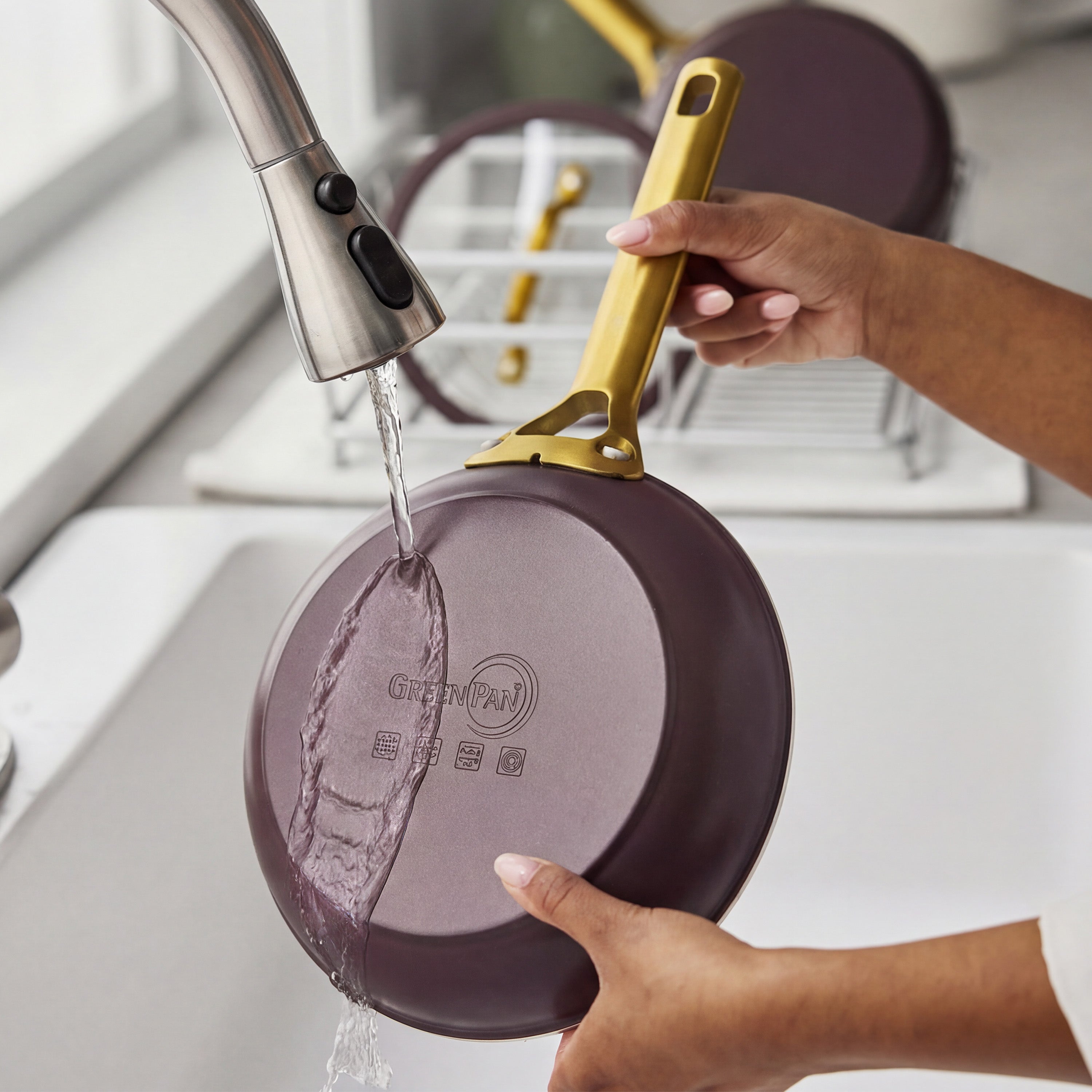 A person rinses a Bordeaux-colored ceramic nonstick pan from a 10-piece cookware set under a kitchen faucet.