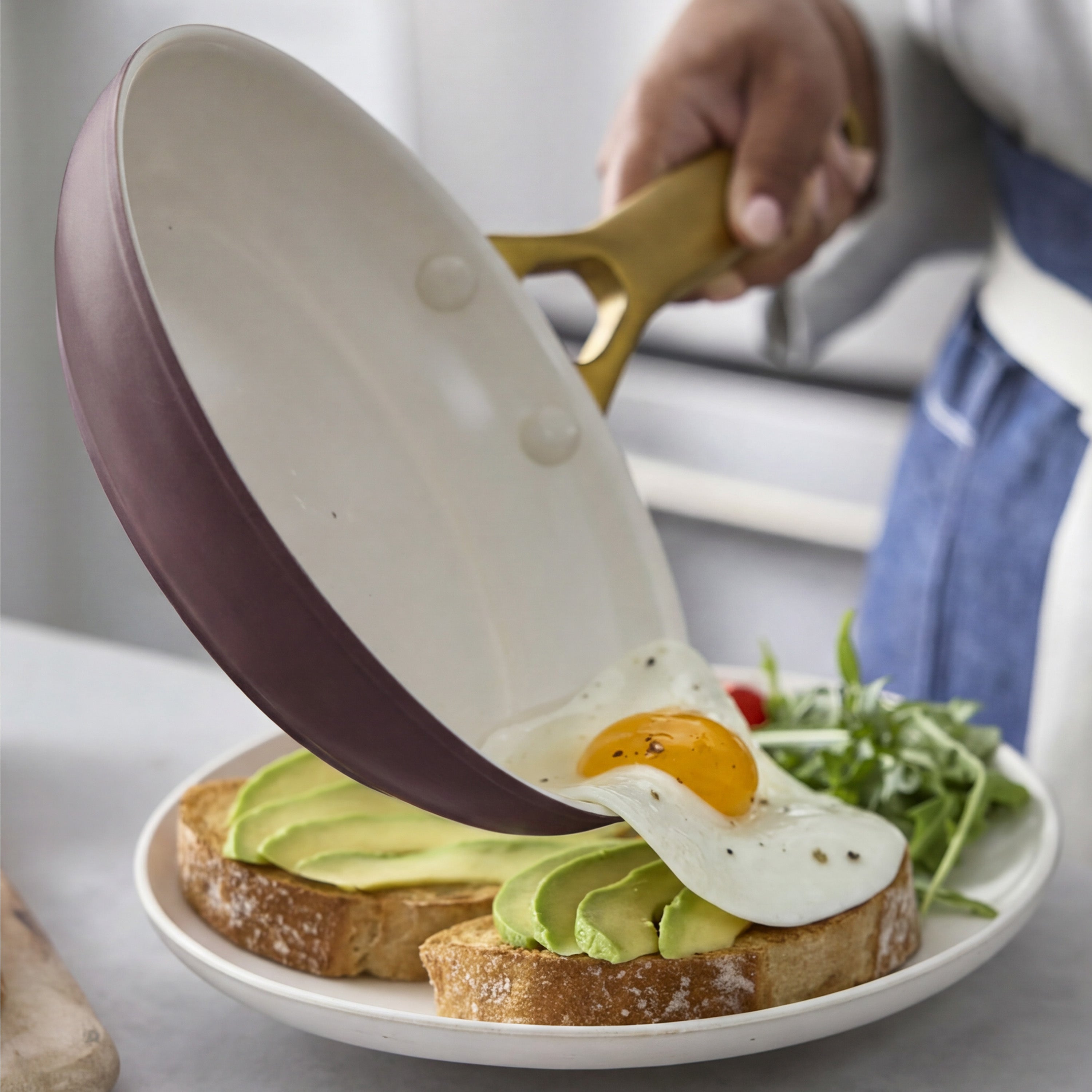 A fried egg slides from a ceramic nonstick pan onto avocado toast with greens on a white plate.