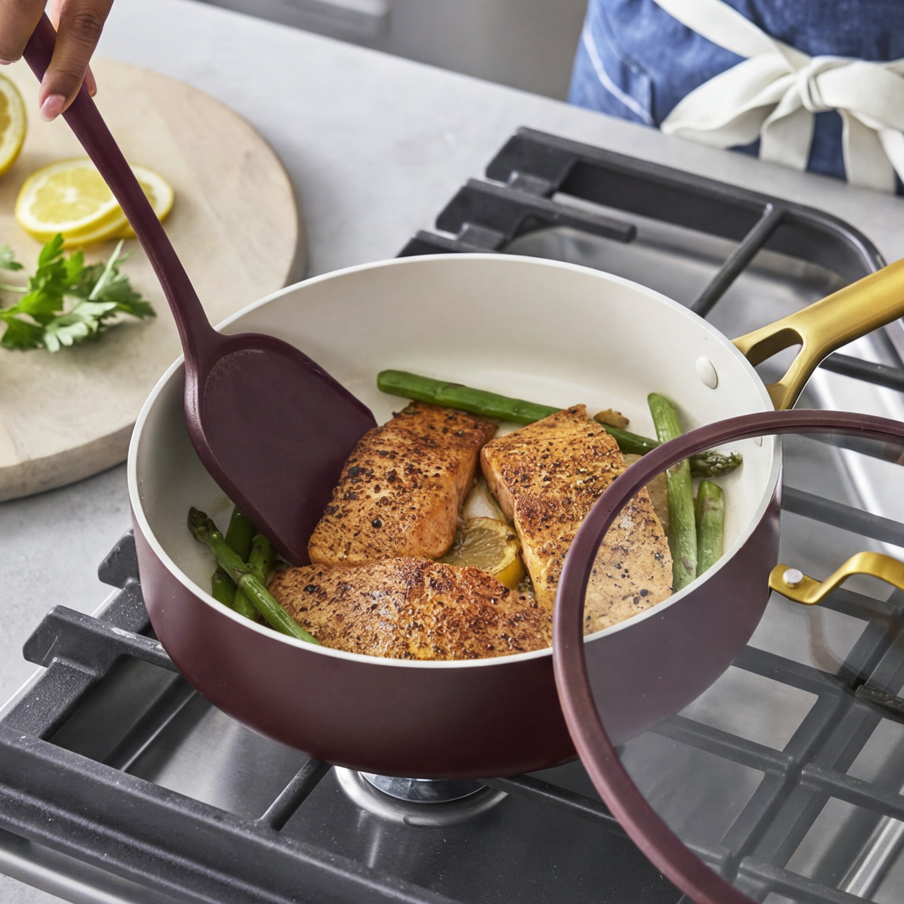 Salmon and asparagus cooking in a Bordeaux ceramic nonstick pan with a spatula, lemon, and parsley nearby.