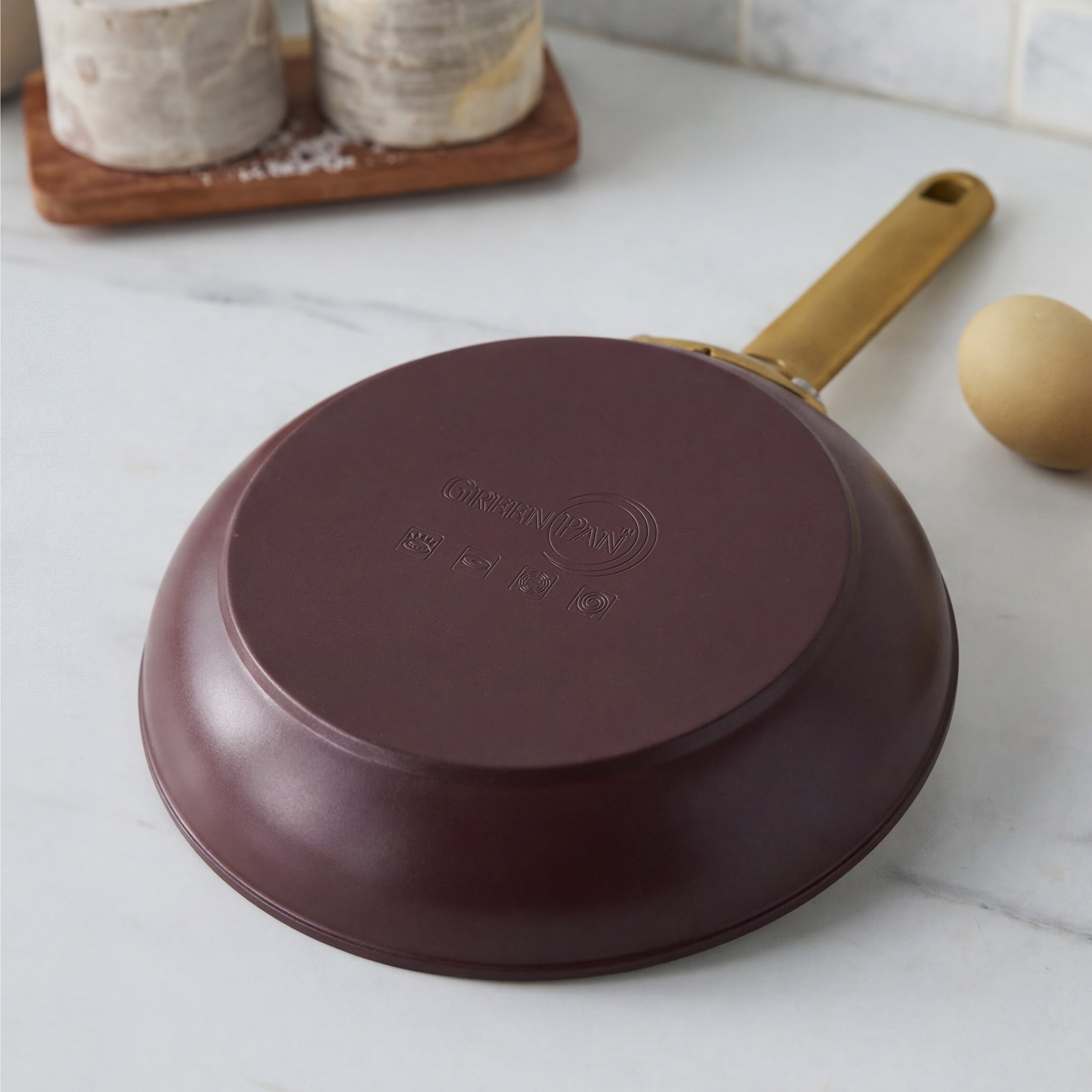 Bordeaux ceramic nonstick pan with gold handle upside down on white counter, next to a brown tray and an egg.