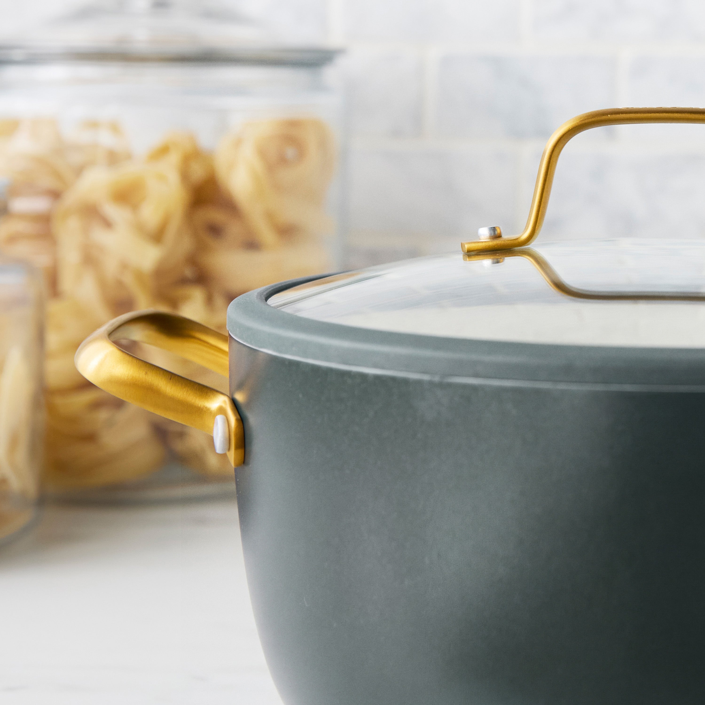 Spruce green ceramic nonstick 10-piece cookware set with gold handles and glass lids, stacked on a kitchen counter near pasta jars.