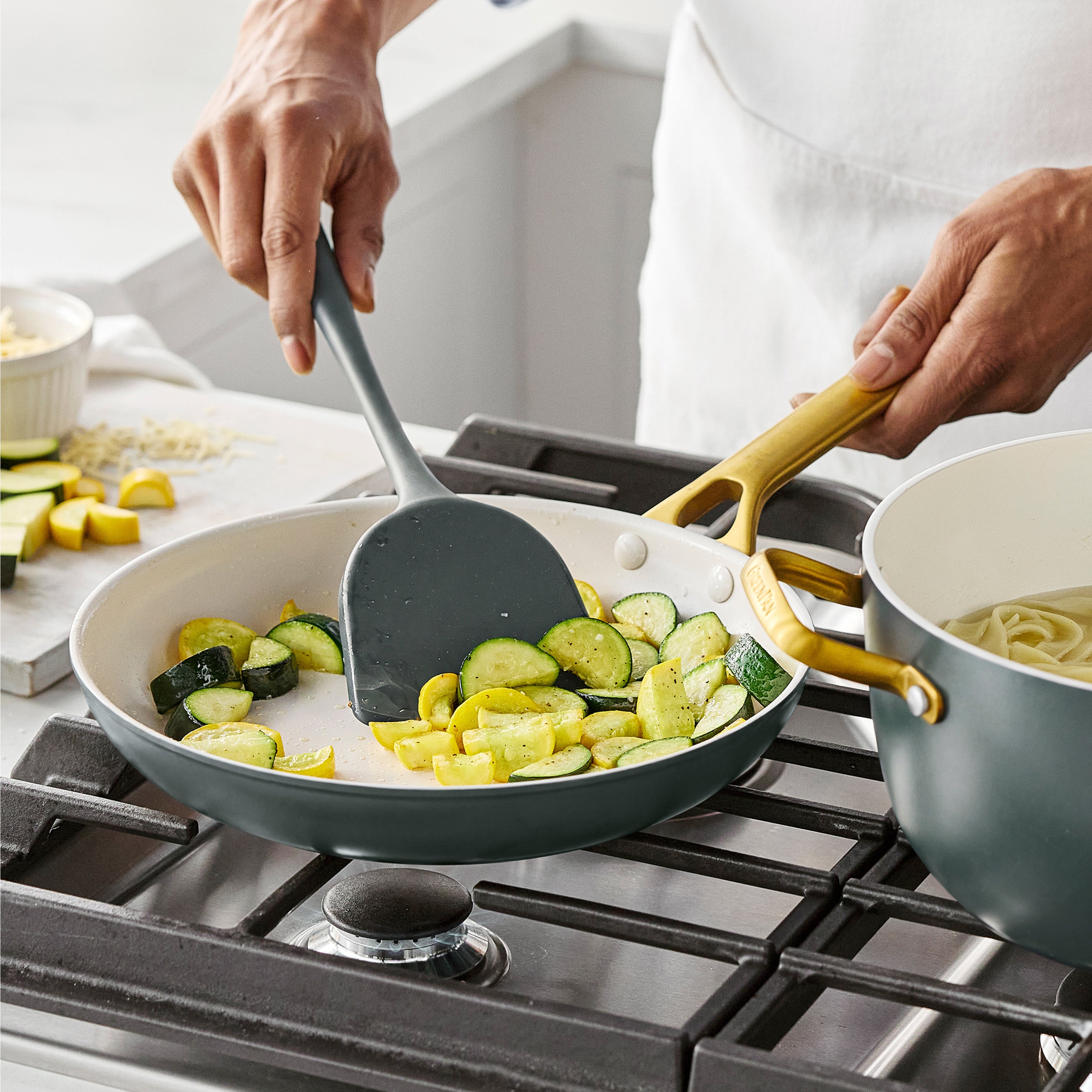 Zucchini and squash cook in a ceramic nonstick skillet; pasta boils nearby, both from a green 10-piece cookware set.