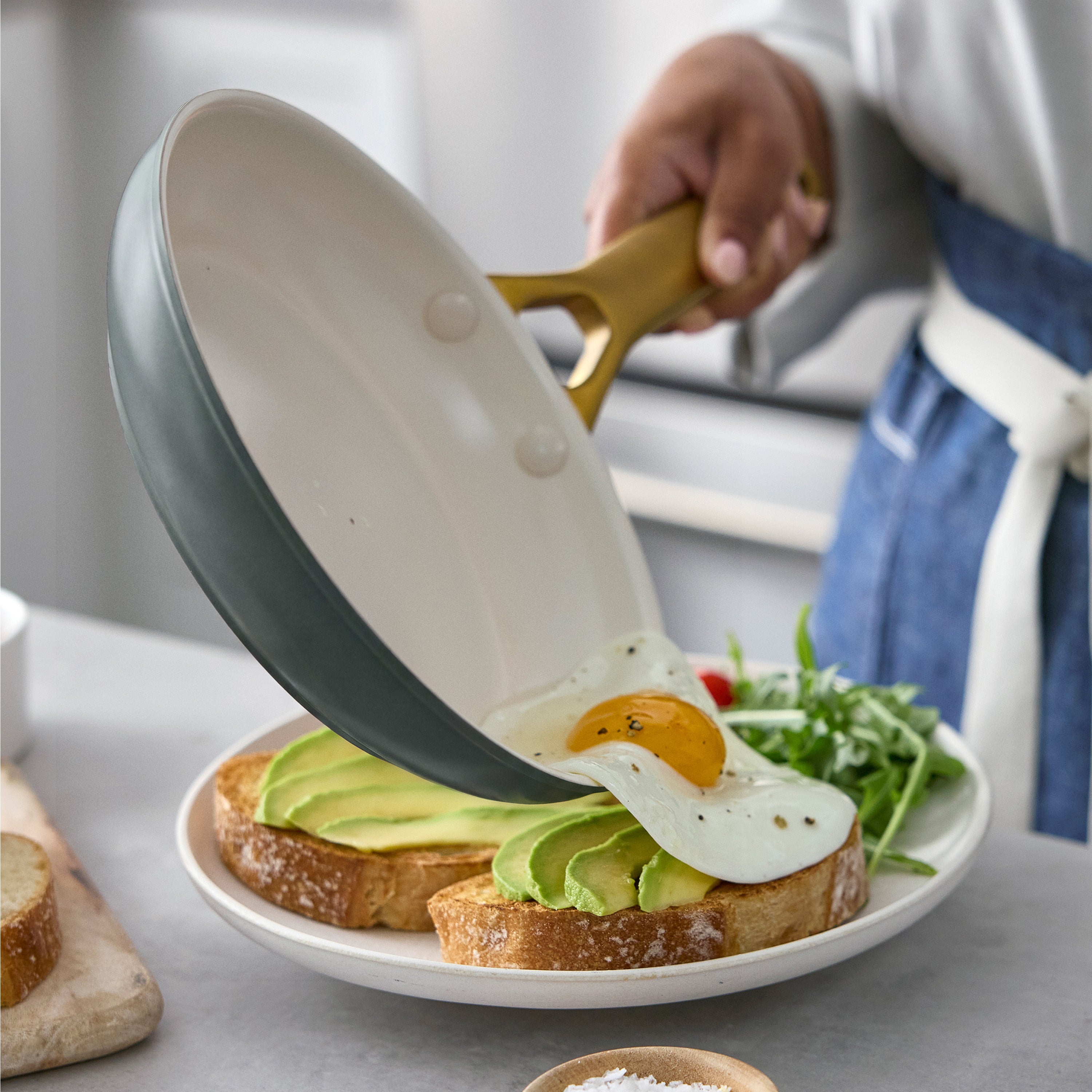 A ceramic nonstick frying pan slides a fried egg onto avocado toast, with greens beside the toast on a light surface.