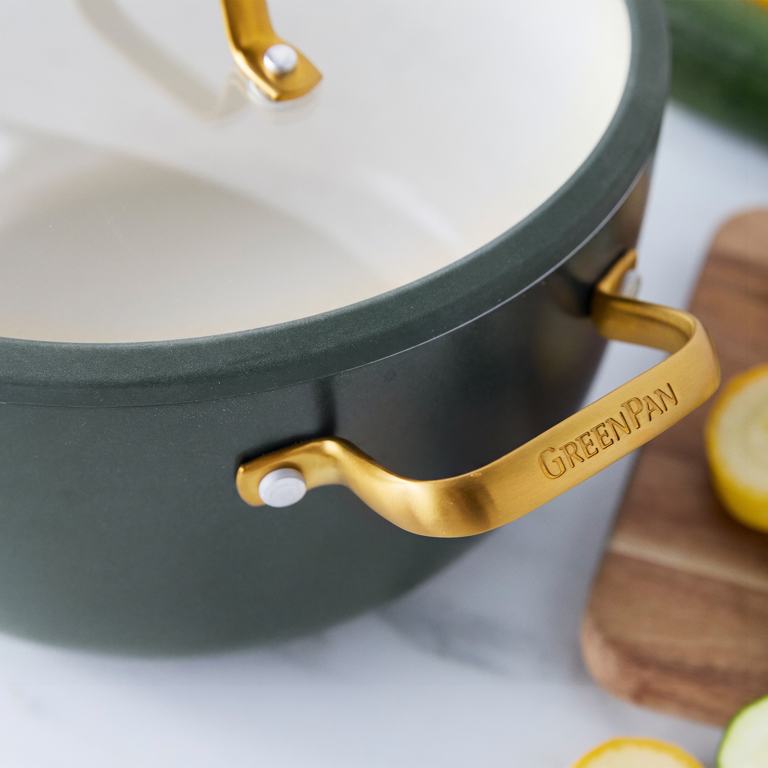 Ceramic nonstick 10-piece cookware set in spruce with gold handles, displayed with sliced vegetables on a cutting board.