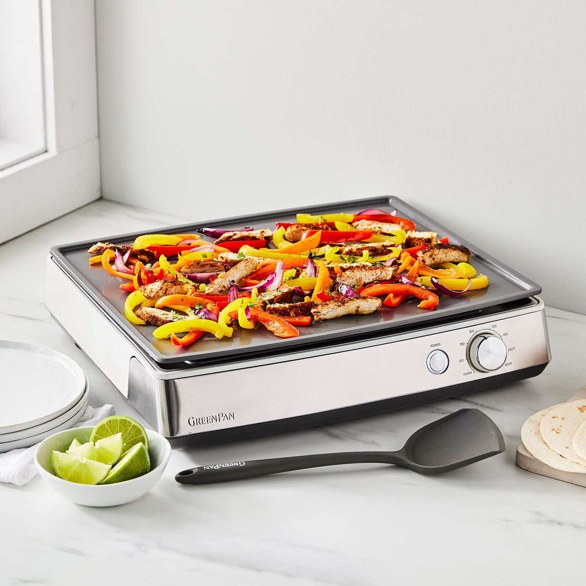 Sliced chicken, peppers, and onions cooking on a Cloud Cream Ultimate Countertop Griddle with tortillas and lime wedges.