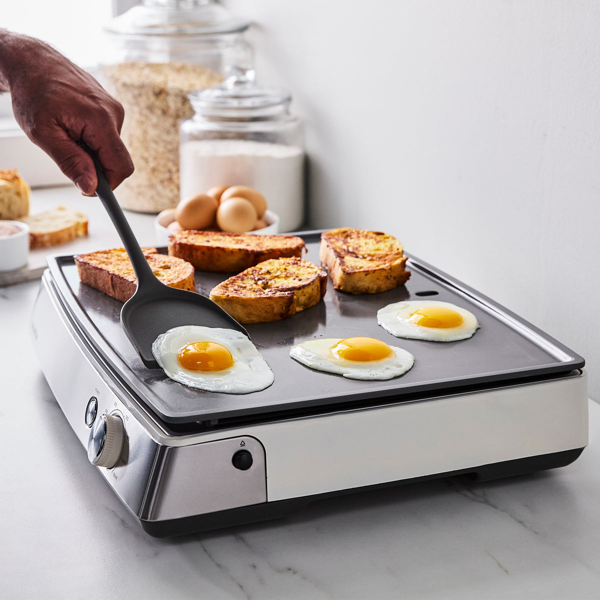 A hand flips a fried egg with a spatula on a Cloud Cream Ultimate Countertop Griddle with French toast cooking nearby.