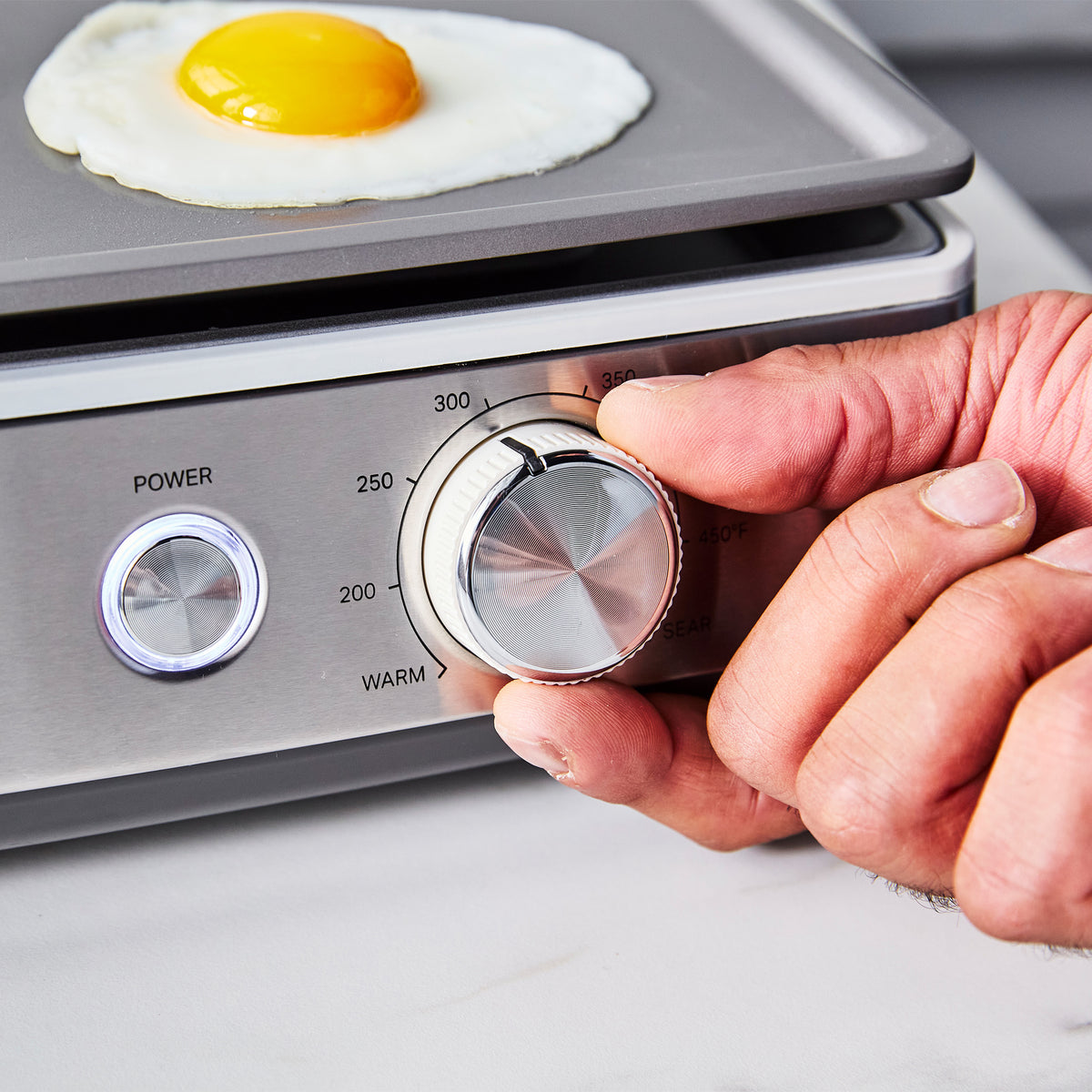 A hand adjusting the temperature dial on a Cloud Cream Ultimate Countertop Griddle while a fried egg cooks on the nonstick surface.