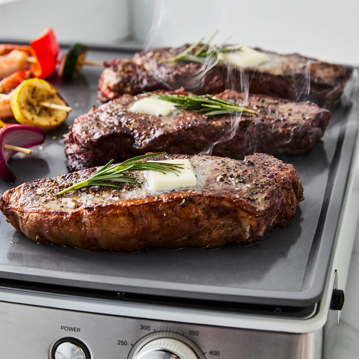 Three steaks searing on an Ultimate Countertop Griddle in Cloud Cream, topped with melting butter and rosemary.