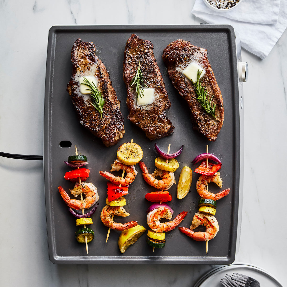 Three steaks, rosemary, butter, and shrimp skewers cooking on a Cloud Cream Ultimate Countertop Griddle.