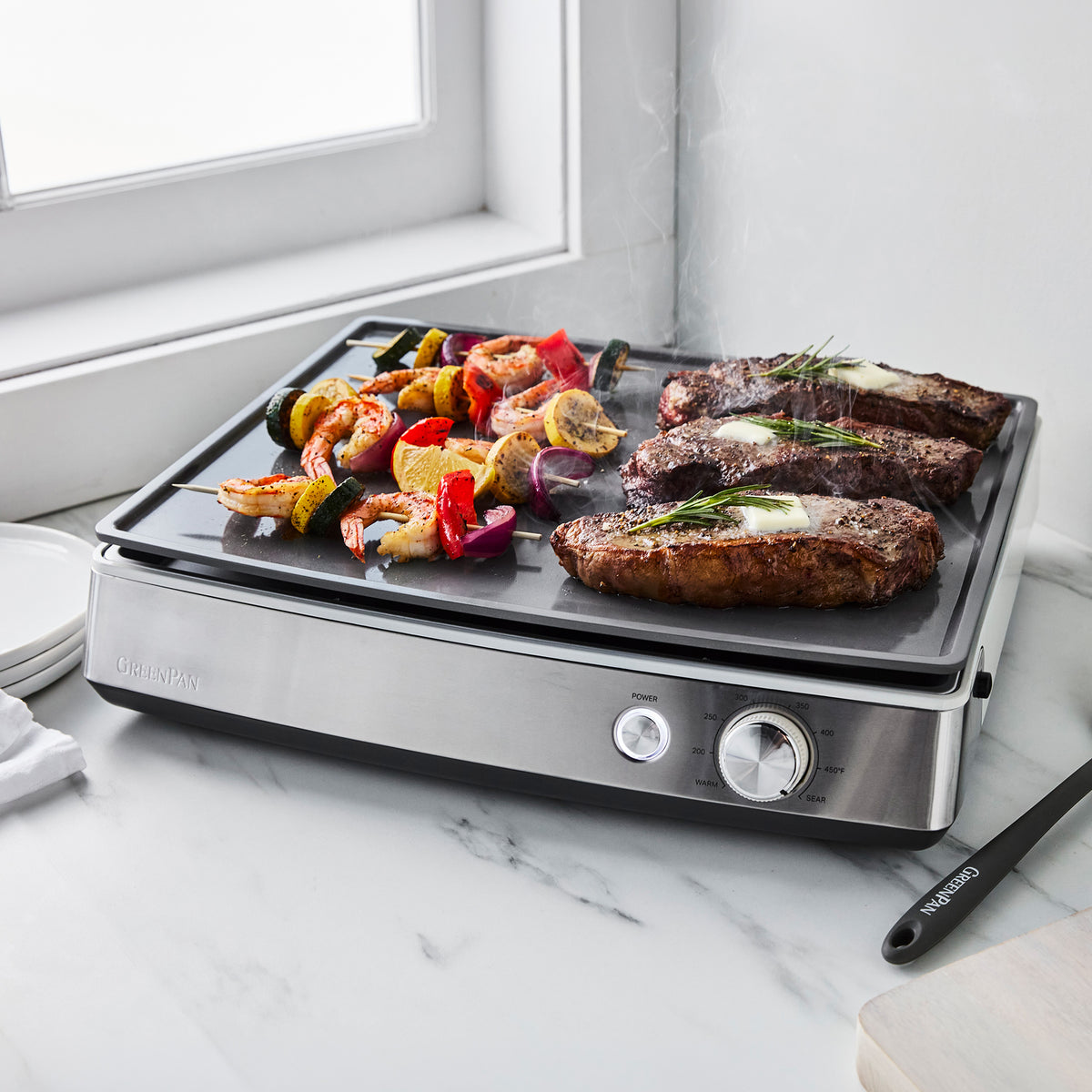 Cloud Cream countertop griddle filled with shrimp skewers, vegetables, and three steaks topped with melting butter.