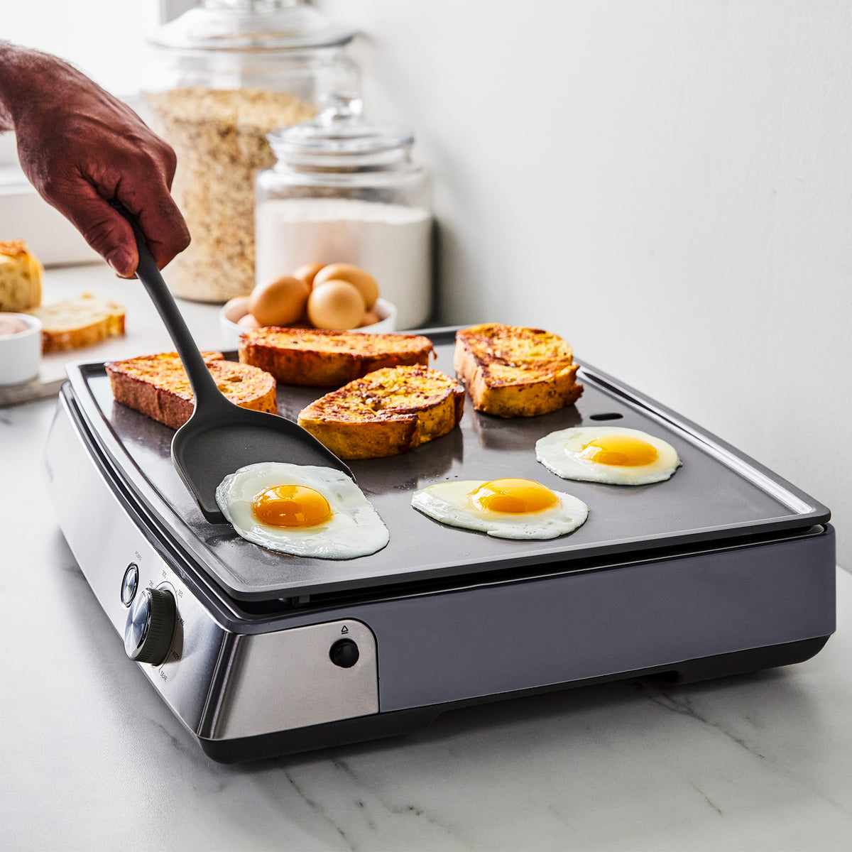 A spatula flips a fried egg on a graphite Ultimate Countertop Griddle alongside two eggs and three slices of French toast.