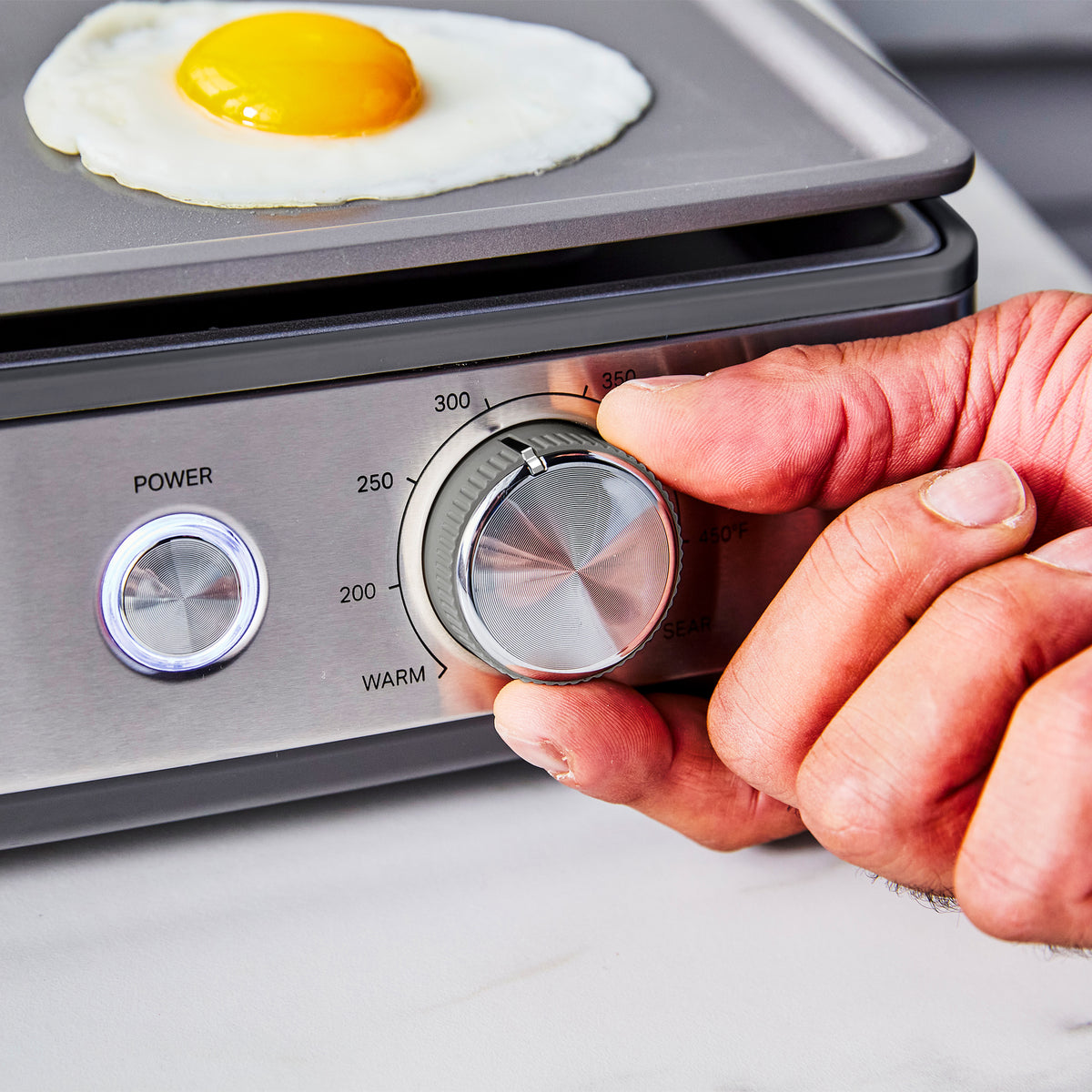 A hand adjusts the temperature dial on a Graphite Ultimate Countertop Griddle as an egg cooks on the nonstick surface.