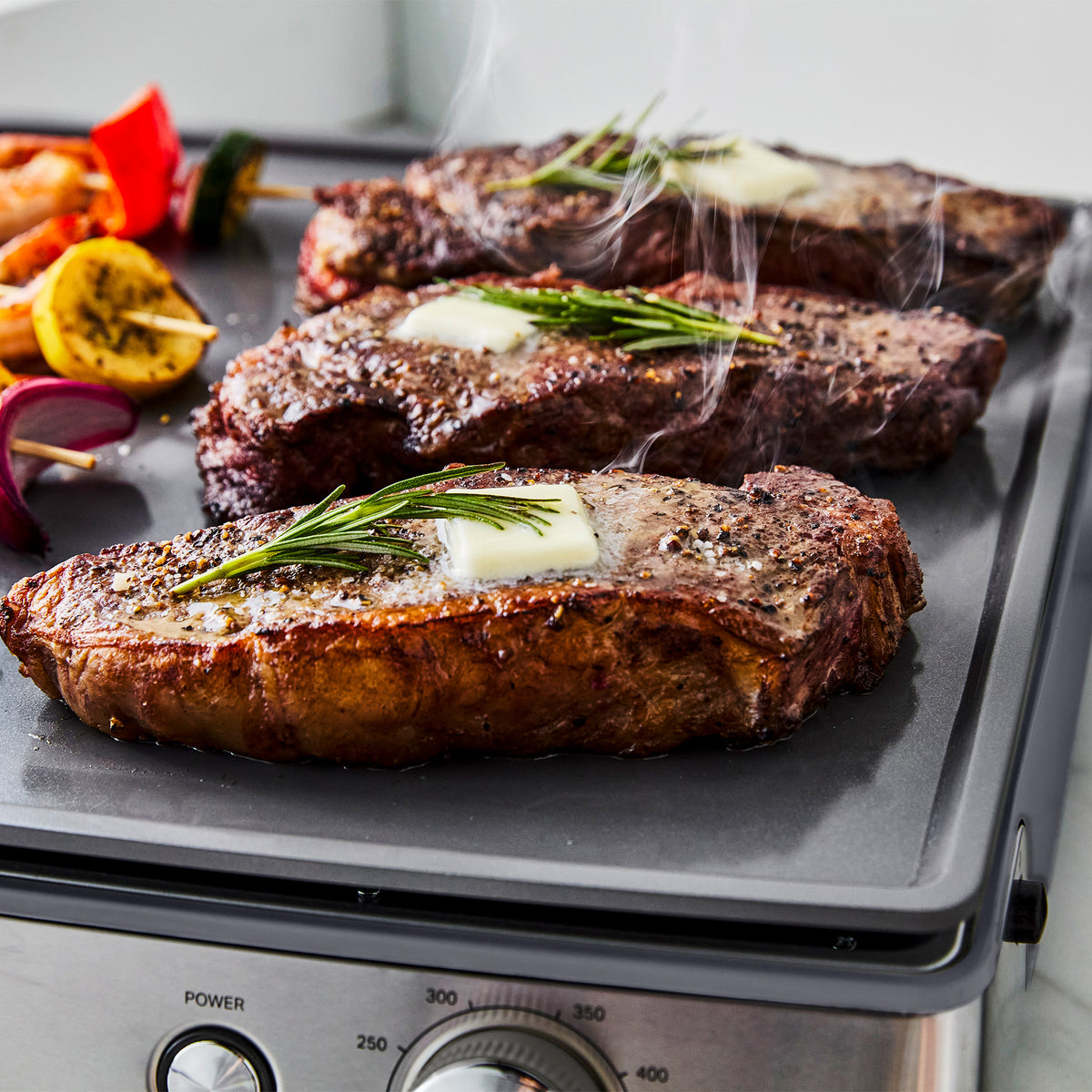 Three steaks topped with butter and rosemary cooking on a graphite Ultimate Countertop Griddle.