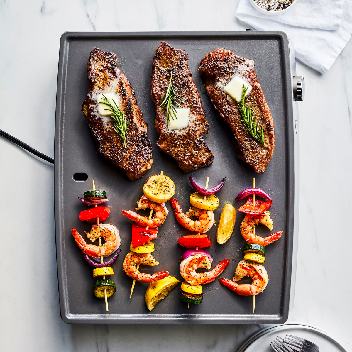 Three seared steaks with butter, rosemary, shrimp, and vegetable skewers on a graphite Ultimate Countertop Griddle.