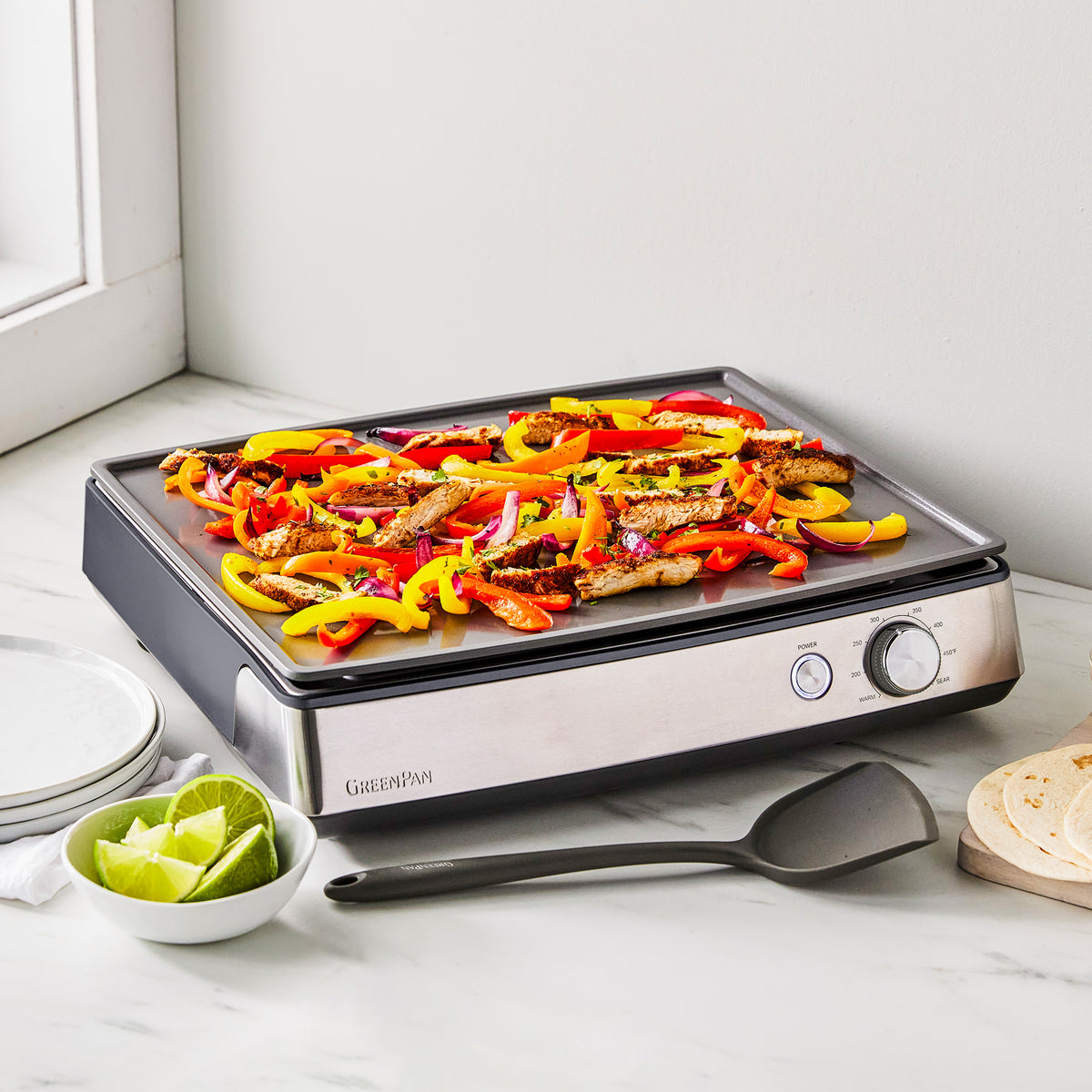 Ultimate Countertop Griddle in graphite cooking sliced chicken, peppers, and onions, with lime wedges and tortillas on the side.
