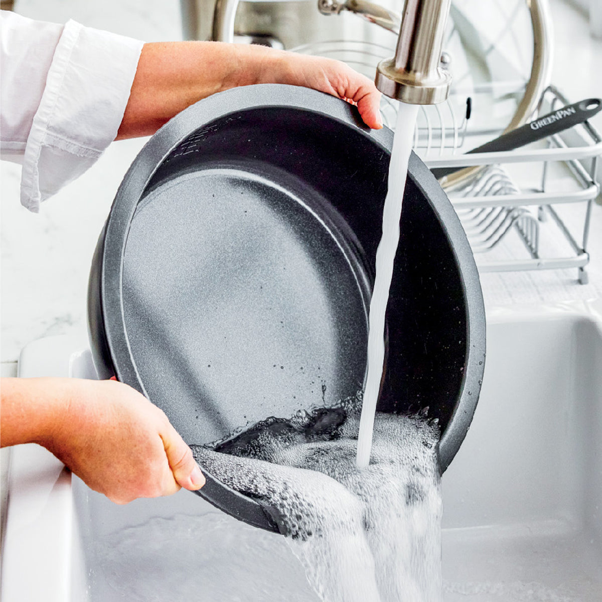 Hand washing a pan with soap under running water; dish rack and utensils visible in the background.