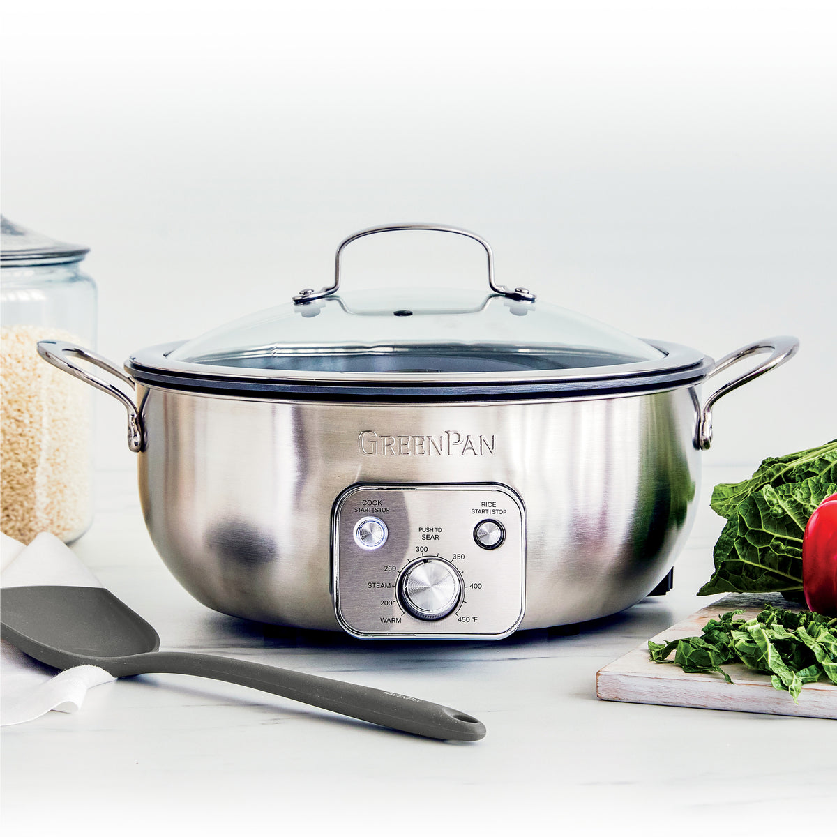 Electric wok with black exterior on a kitchen counter, next to assorted vegetables, grains, and a spatula.