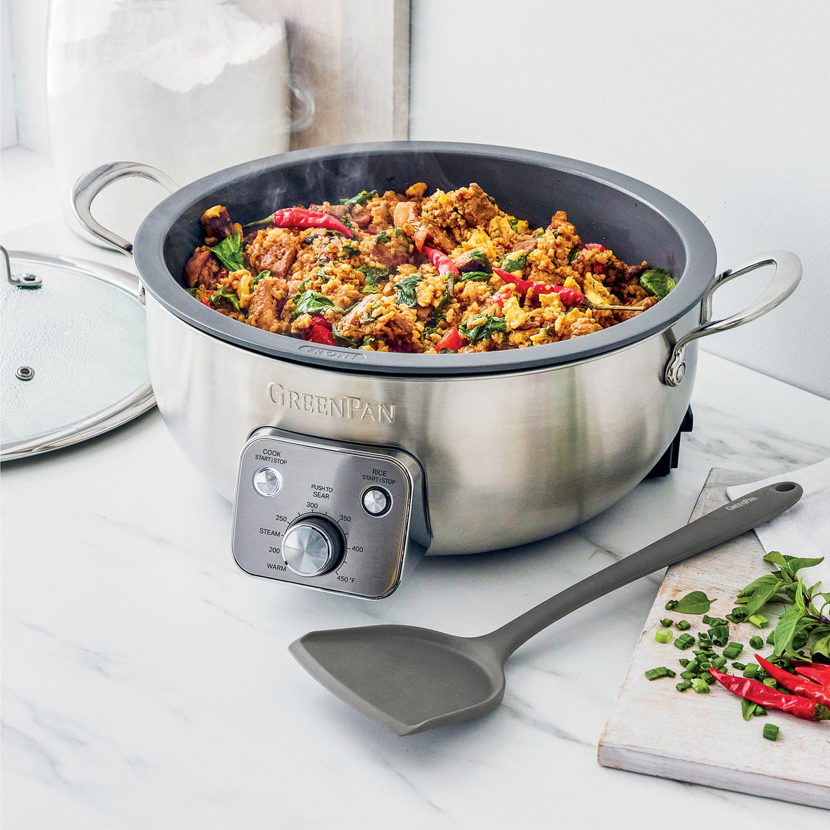 Electric wok with meat and vegetables, a spatula inside, on a white countertop.