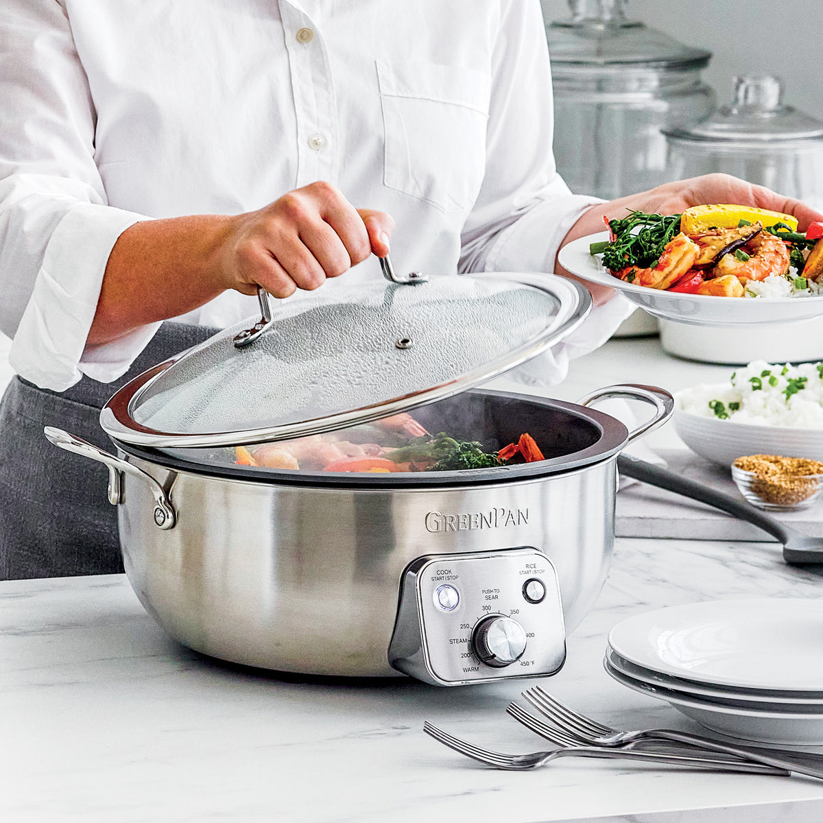 A person lifts the lid of a black Premiere 6-quart electric wok on a counter with stacked white dishes and a plate of food.