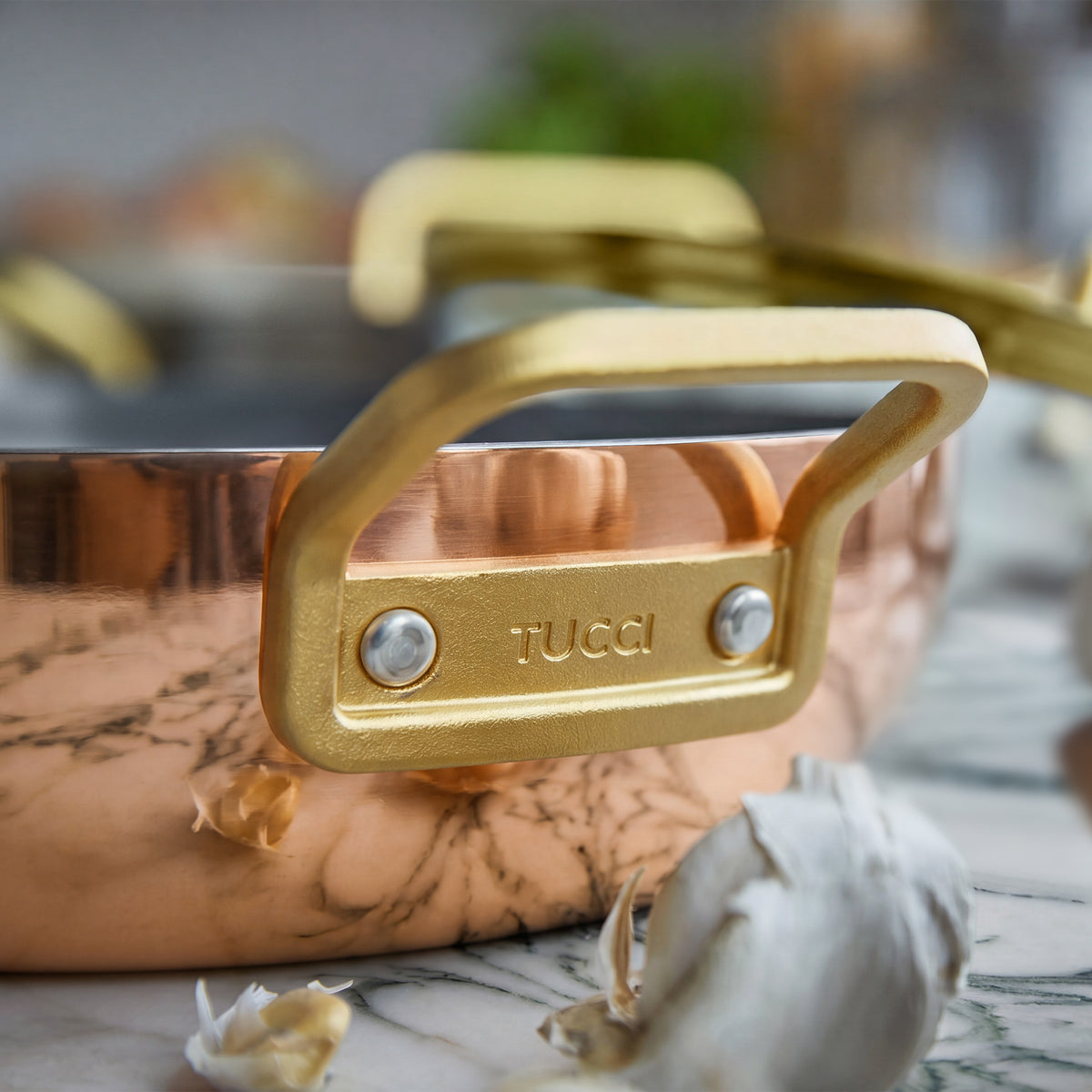 Copper 4.5-quart saucepan with gold handles next to garlic cloves on a marble surface.
