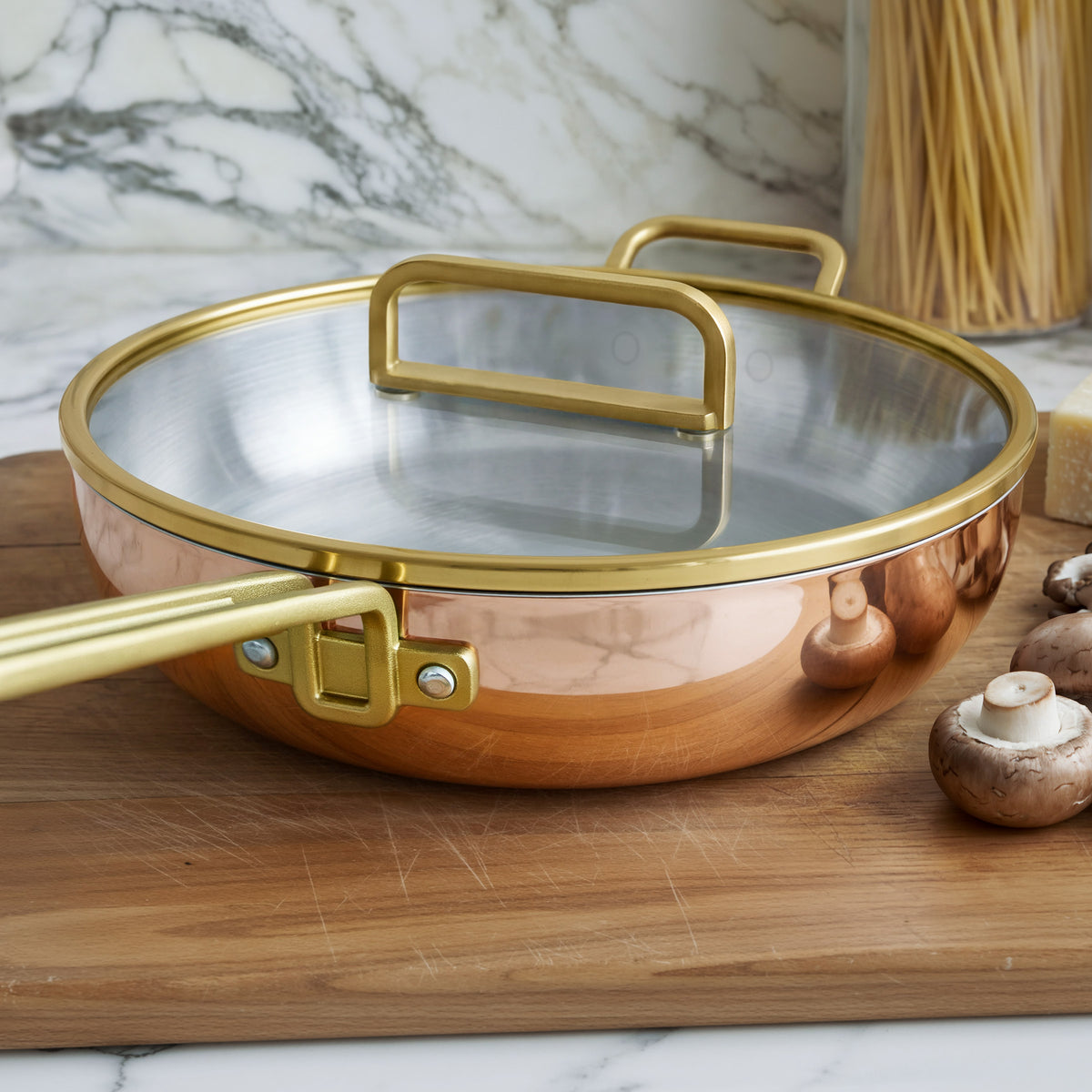 Copper pan with gold handles and lid on a board, surrounded by mushrooms and pasta.