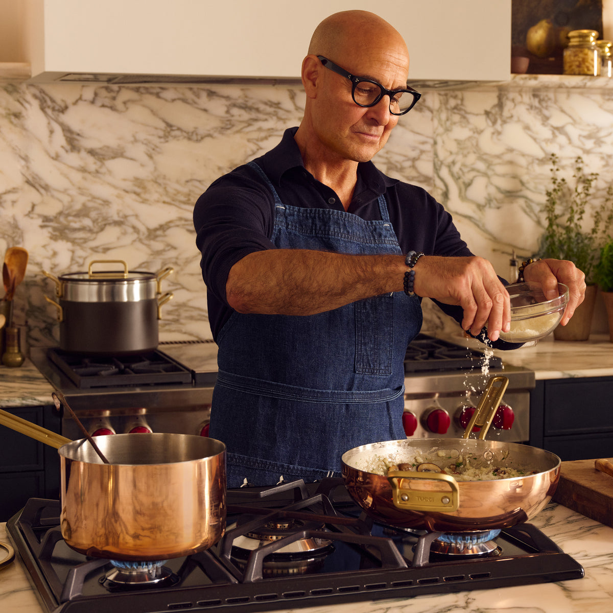 Man wearing glasses and apron seasons food in a copper Stanley Tucci™ Copperply 4.5-quart pan on a modern kitchen stovetop.