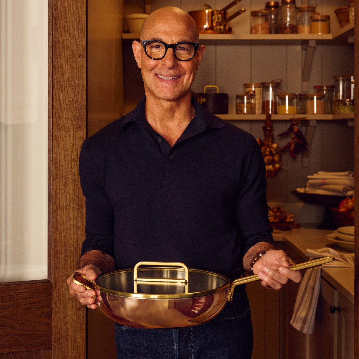 Man wearing glasses holding a copper Stanley Tucci 4.5-quart saucepan in a warm kitchen with shelves of glass jars in the background.