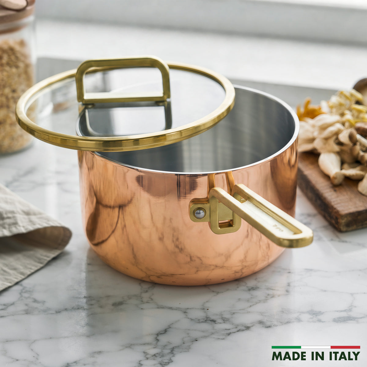 Copper 4-quart saucepan with lid on a marble counter, surrounded by mushrooms and grains. Made in Italy.