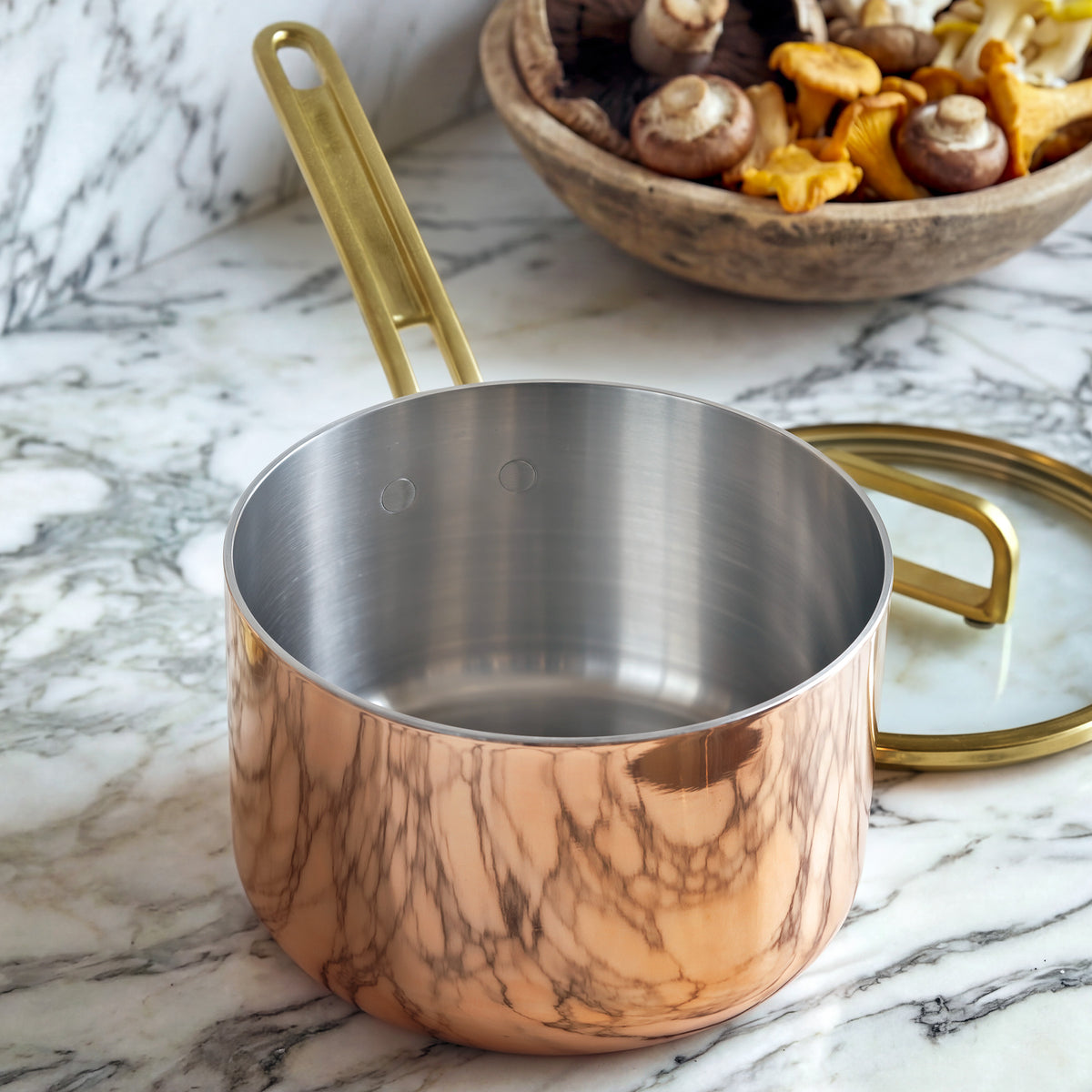 Copper saucepan with gold handle and lid on marble surface, next to a bowl filled with assorted mushrooms.