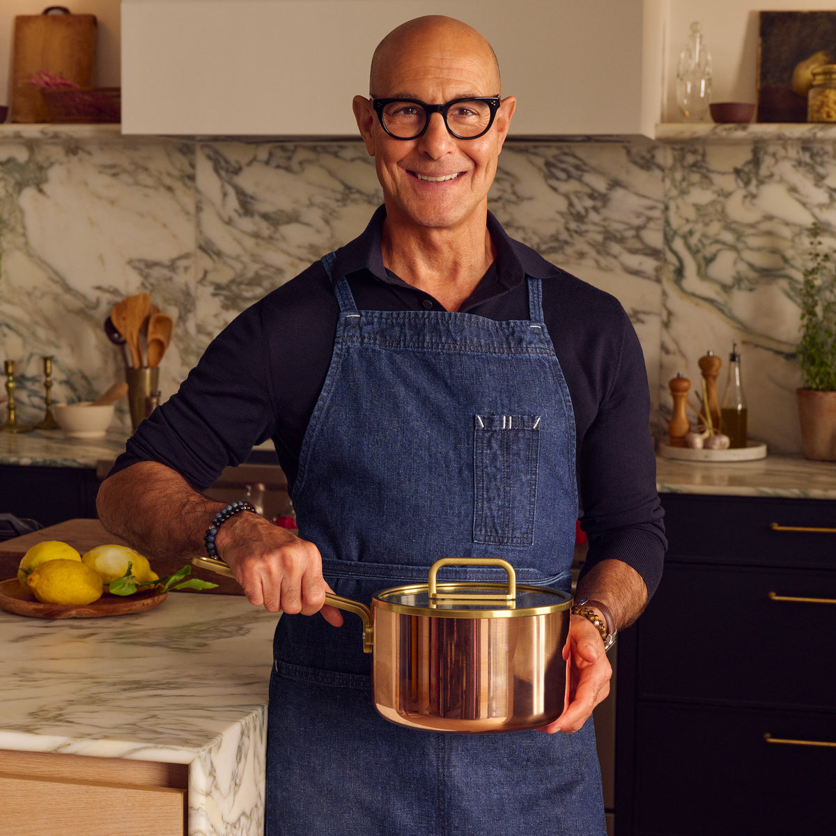 Man in glasses and blue apron holding a copper Stanley Tucci 4-quart saucepan with lid in a modern kitchen.