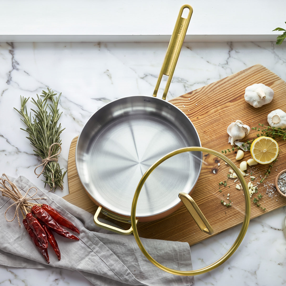Copper sauté pan with gold handle and lid on wood, surrounded by fresh herbs, garlic cloves, lemon slices, and red chilies.