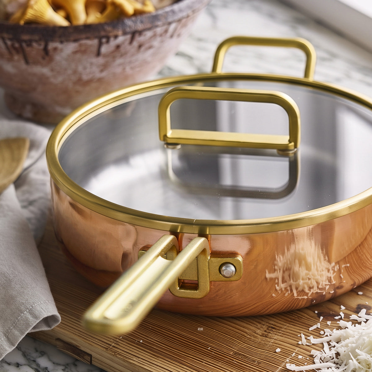 Copper 4-quart sauté pan with lid and gold handle on wood surface, surrounded by shredded cheese.